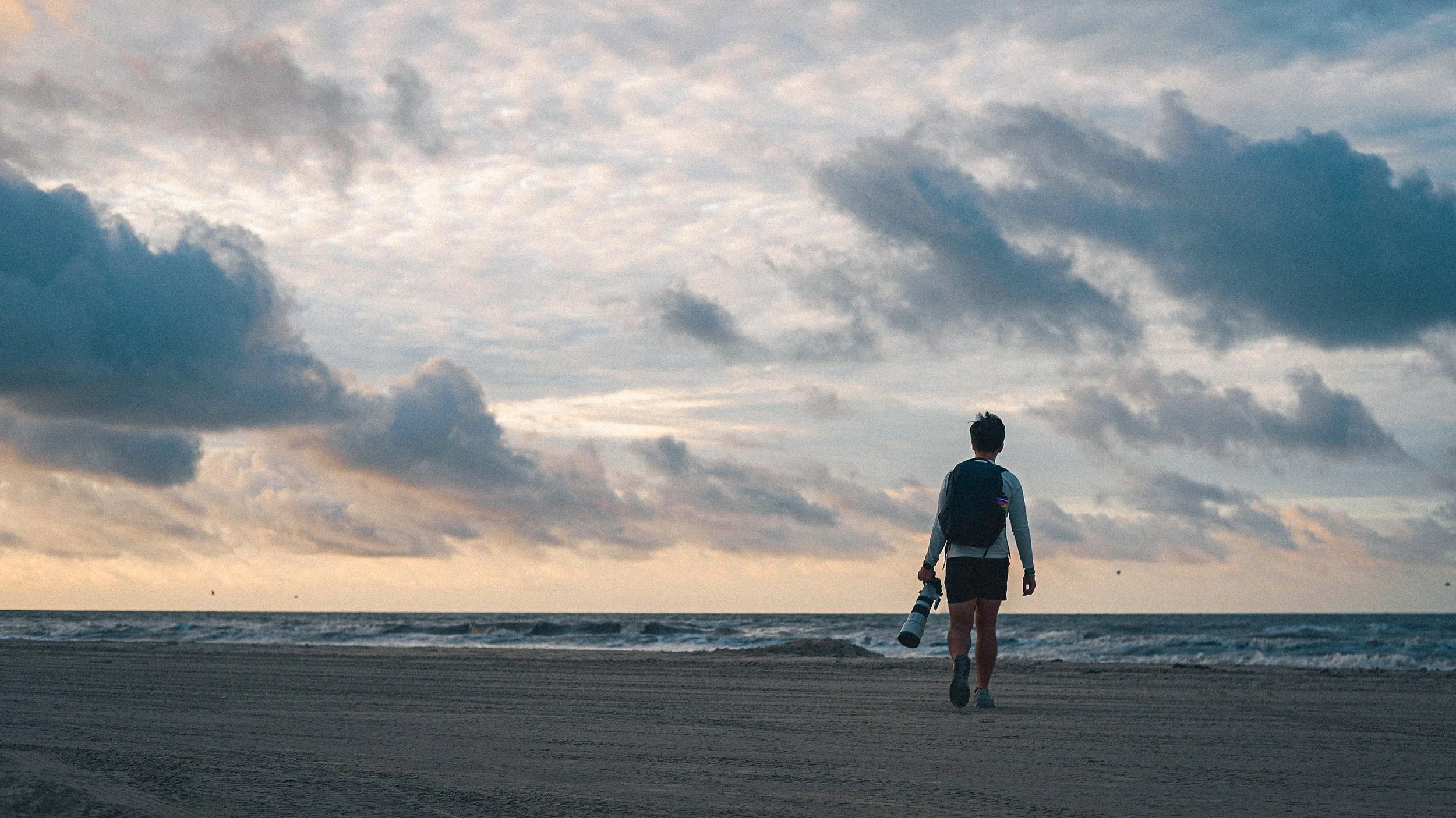 A person with a backpack holding a camera walking on the beach toward the ocean under a cloudy sky.