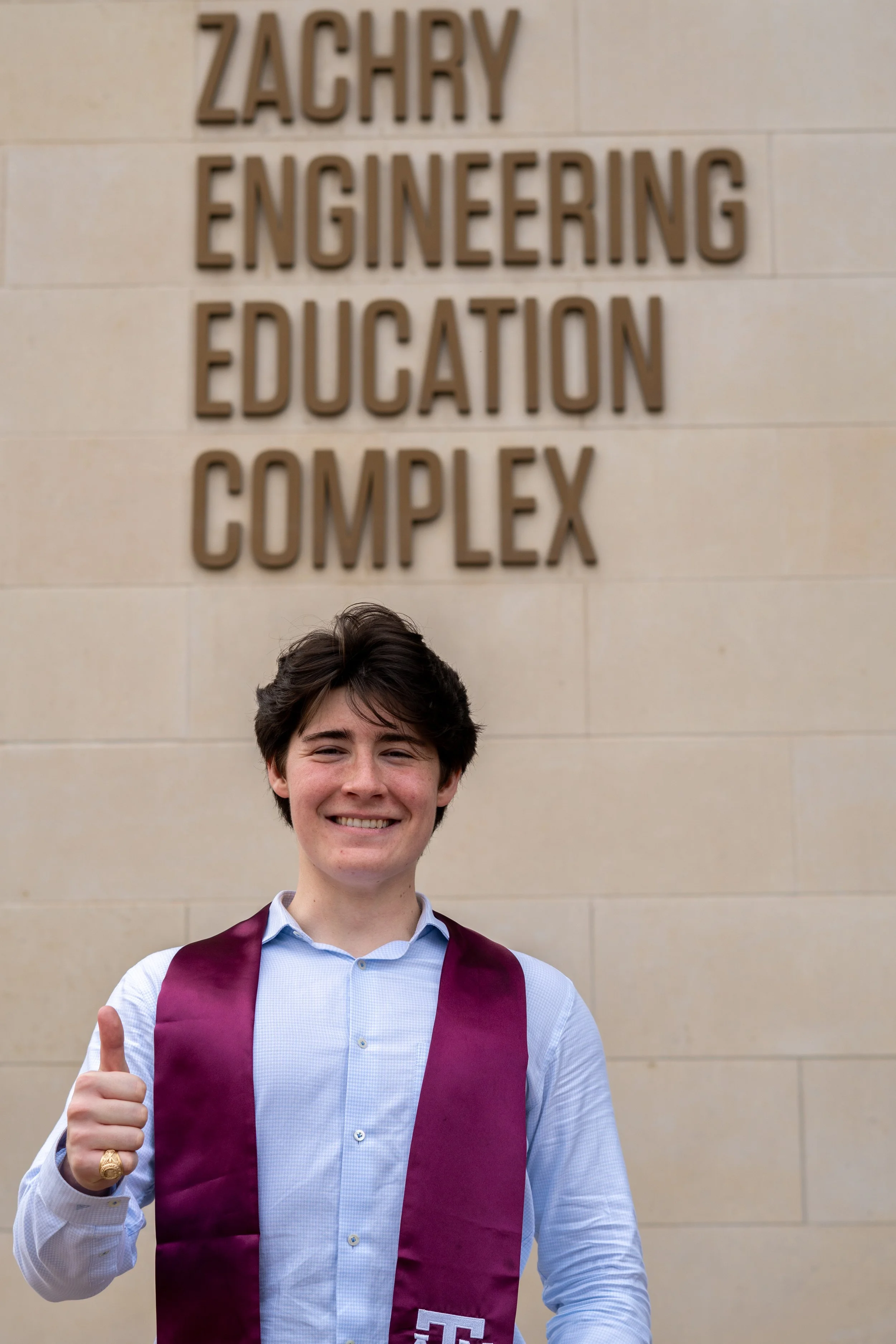 A young man smiling and giving a thumbs-up, dressed in a white shirt and a maroon academic gown with a burgundy sash, standing in front of a sign that reads 'Zachry Engineering Education Complex'.