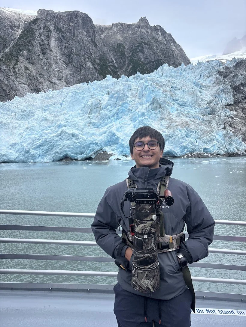 A young man smiling on a boat with a glacier and mountains in the background.