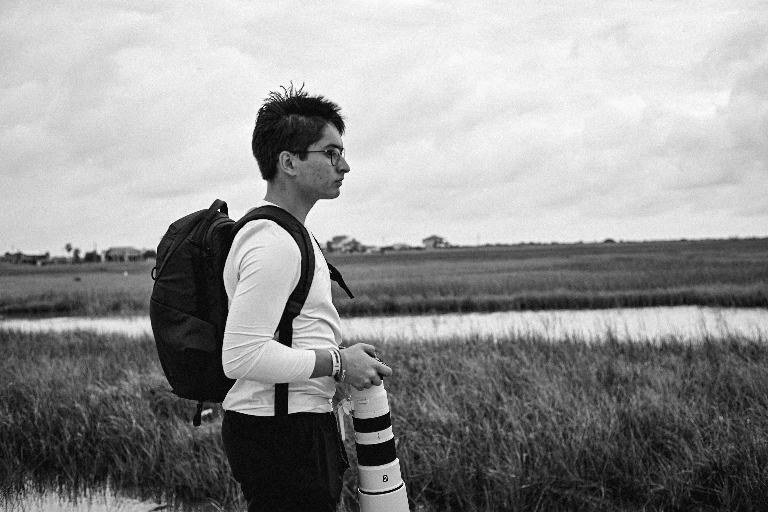 A young man with short spiked hair, glasses, and wearing a white long sleeve shirt with a backpack, standing outdoors in a rural area with fields and a cloudy sky, holding a camera.