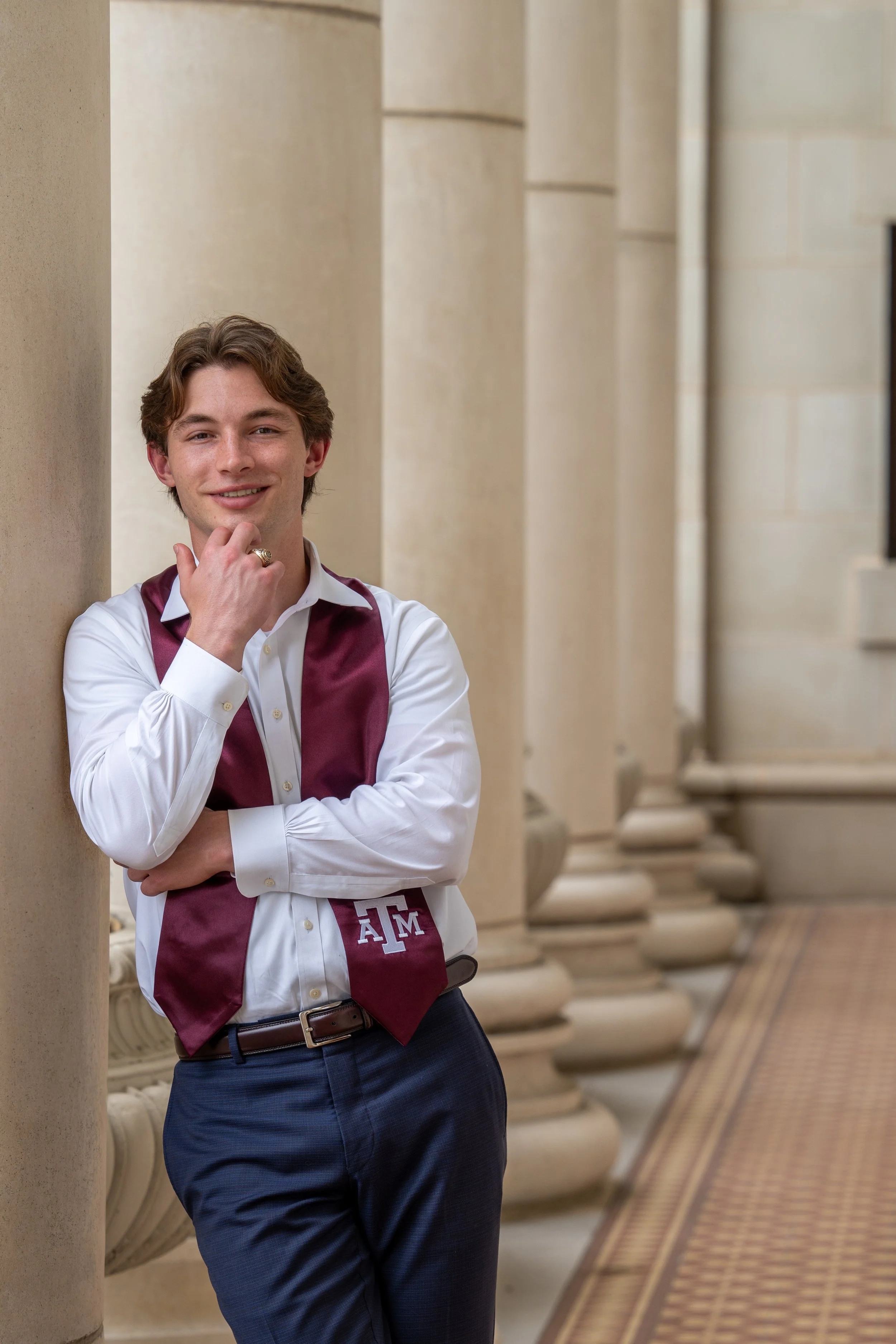 A young man wearing a white shirt, a maroon vest with 'A&M' on it, and navy blue pants, standing outdoors near a row of stone columns.