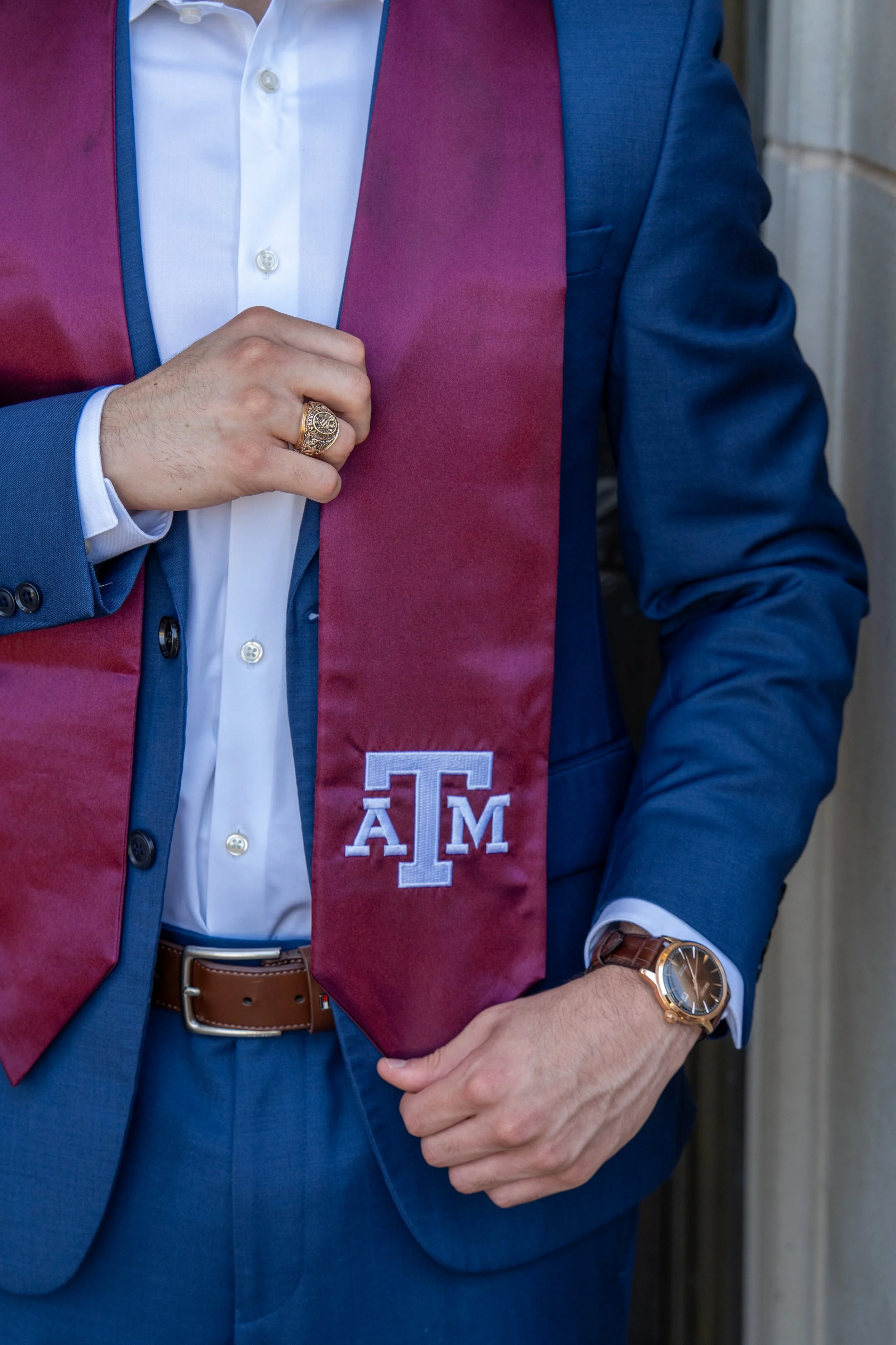 A man in a blue suit with a white shirt and gold watch, wearing a maroon and blue graduation stole with the Texas A&M University logo, standing against a neutral background.