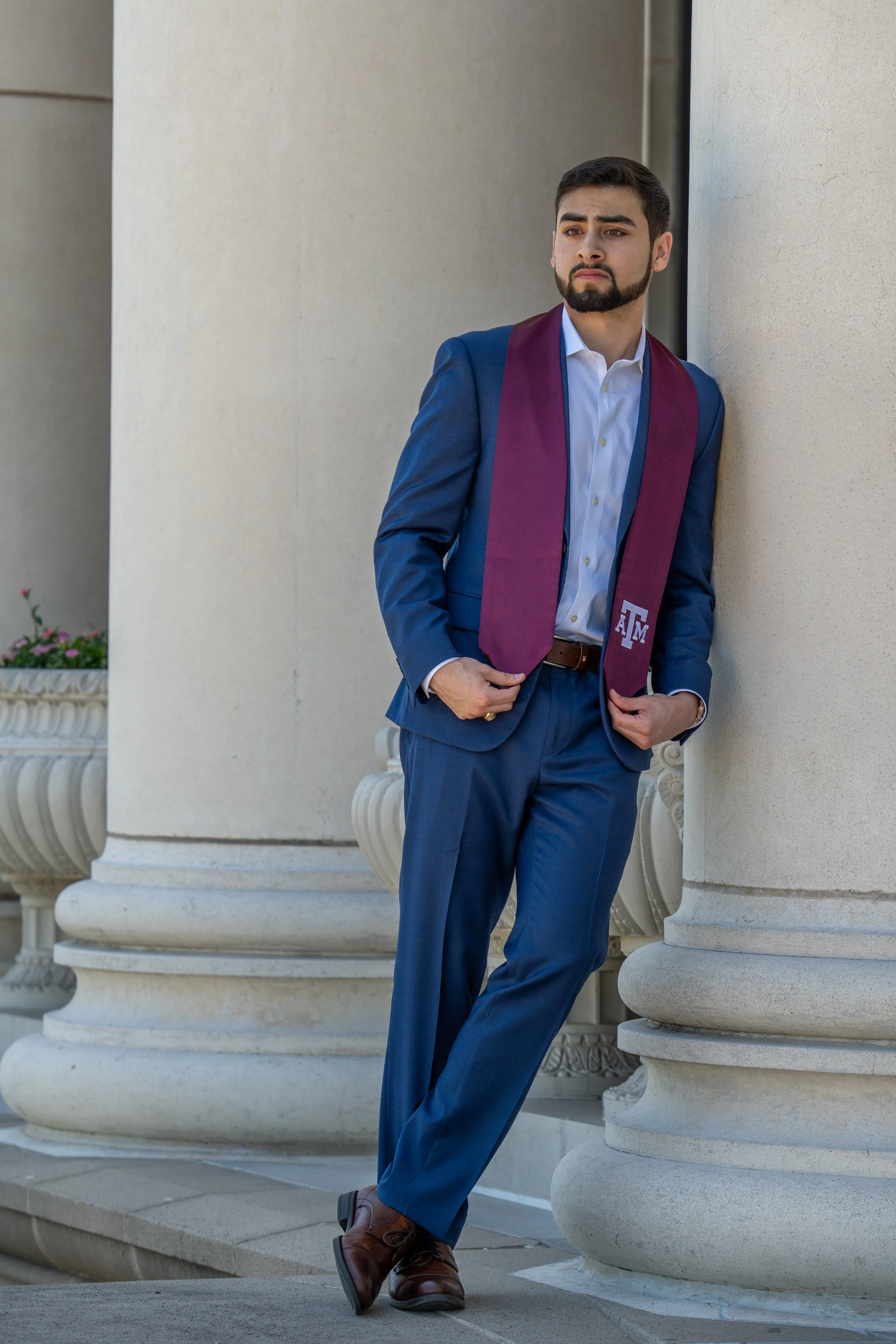 A young man dressed in a blue suit with a maroon stole stands leaning against a large stone pillar outside a building with classical architectural details. He is looking thoughtfully into the distance, with one hand in his pocket and the other holding the edge of his suit.