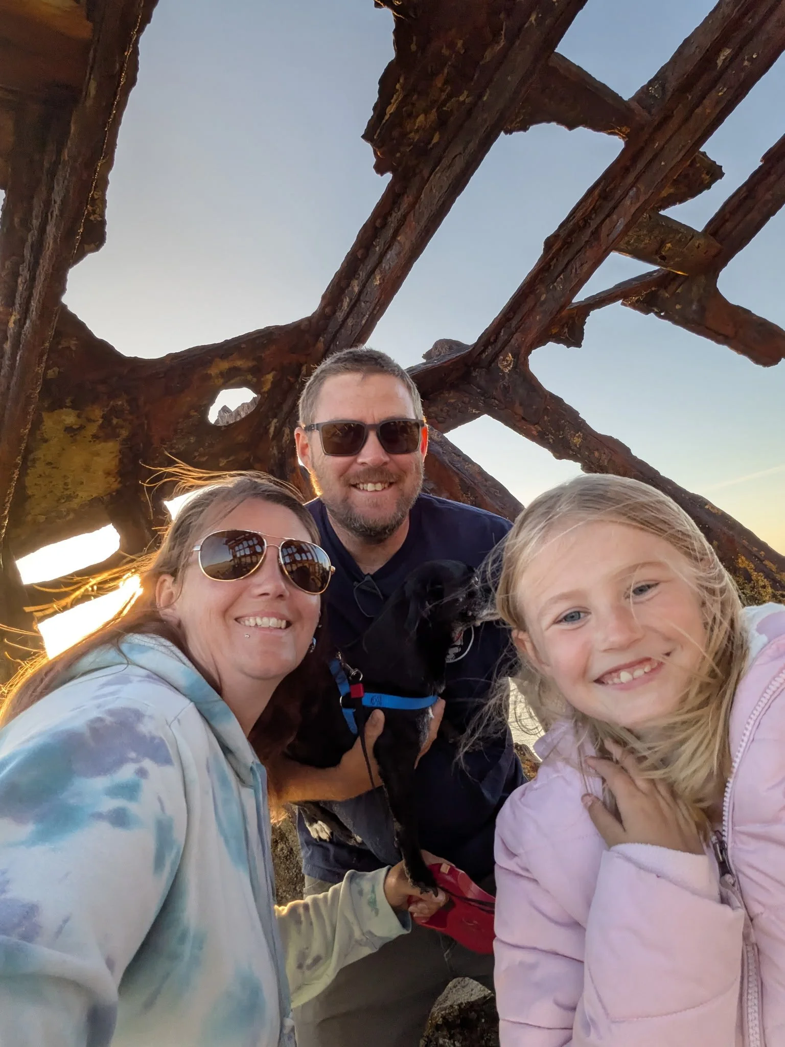 A group of four people smiling, including two women, a man, and a young girl, taking a selfie at a scenic outdoor location during sunset with a large, rusty metal structure behind them.