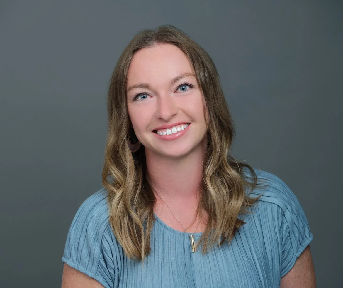 A woman with light brown hair, blue eyes, and a bright smile wearing a blue top, against a plain gray background.