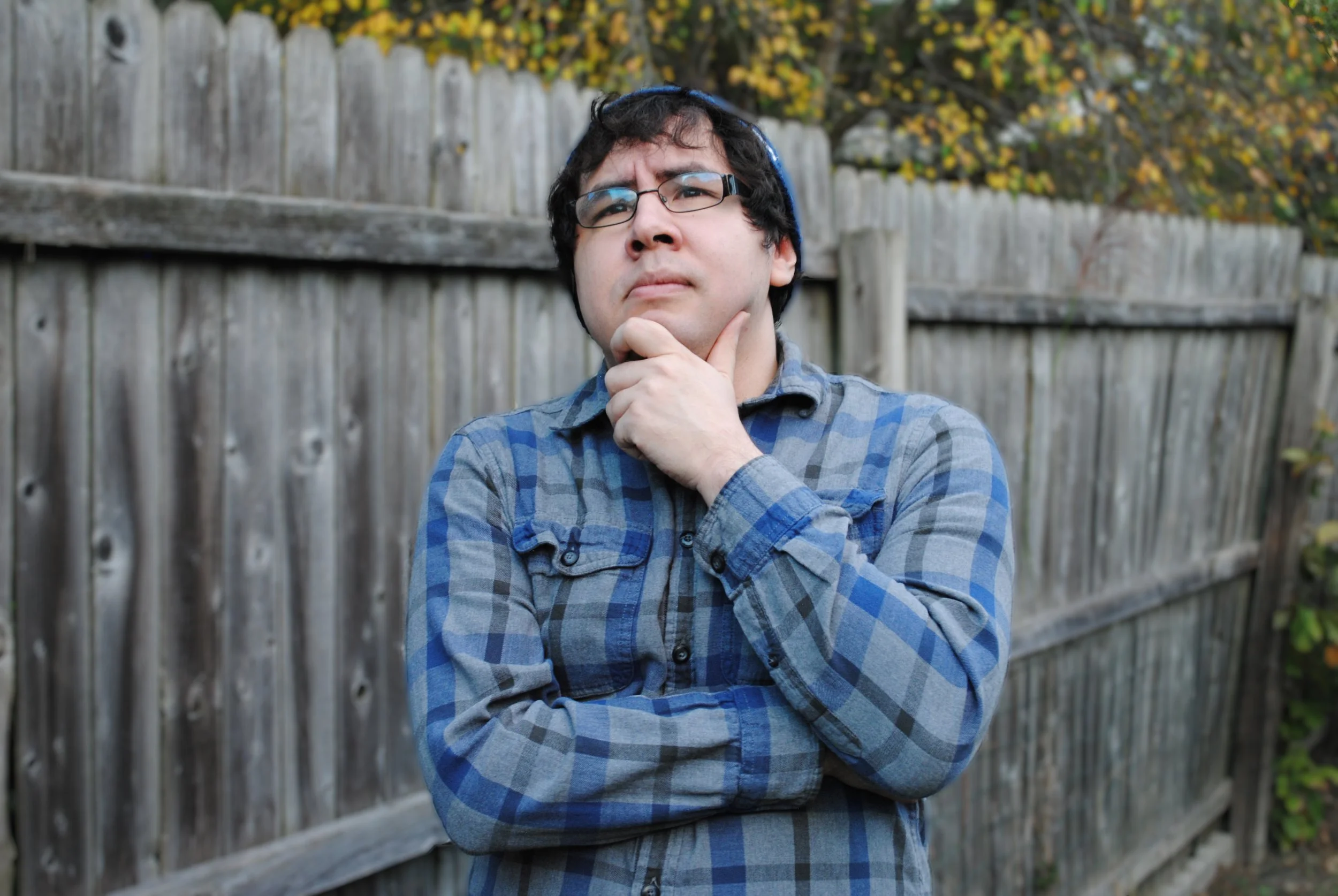 Brian Guerrero with dark hair, wearing glasses and a blue knit hat, stands outdoors with a wooden fence and autumn leaves in the background, holding his chin contemplatively.