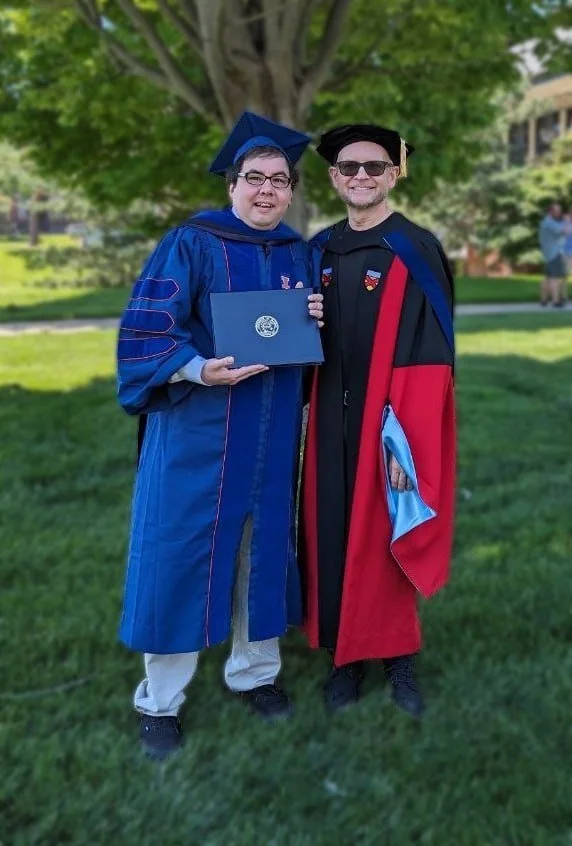 Brian Guerrero in graduation gowns and caps standing outdoors on grass, smiling at the camera. One holds a diploma. There is a large tree behind them.