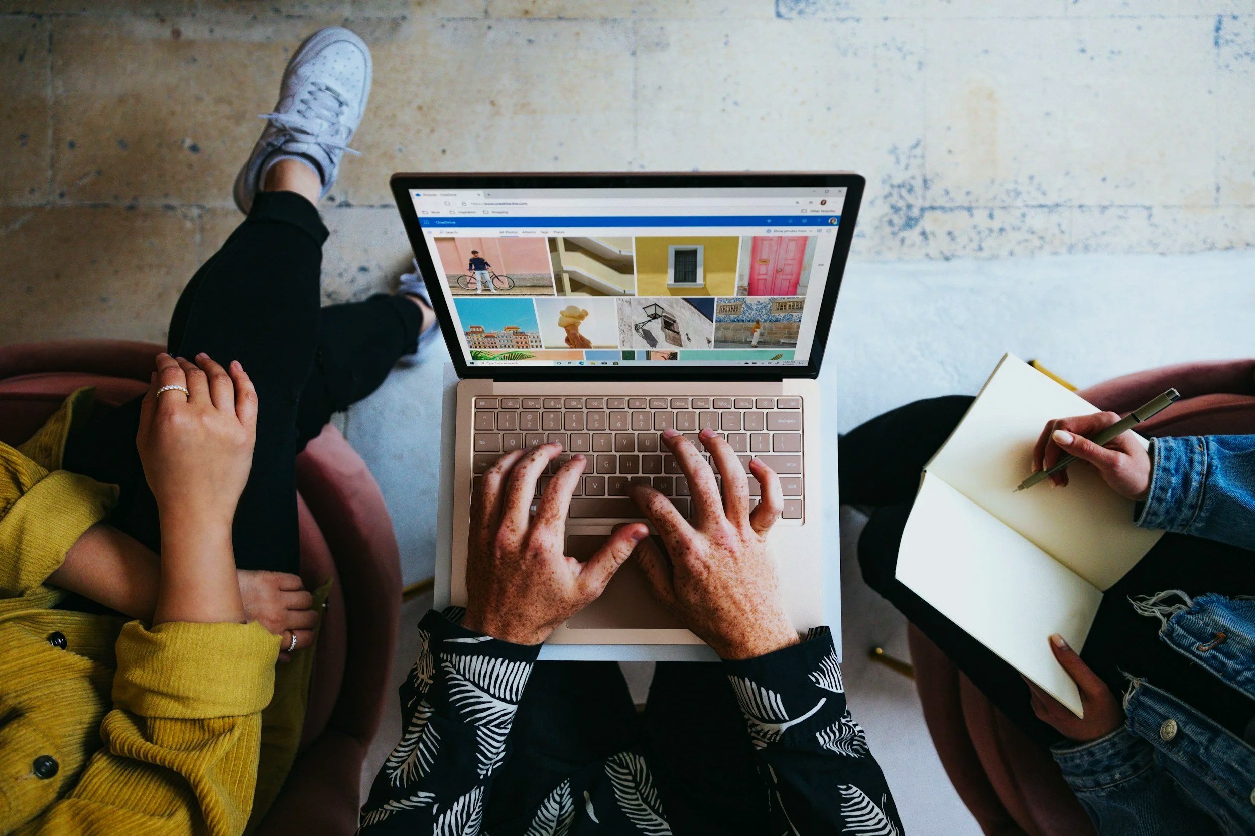 Two people are sitting at a table with a laptop open displaying a photo gallery, and a third person is working on a notepad with a pen. One person is wearing a yellow top, another has animal print sleeves, and the third is in a blue denim jacket.
