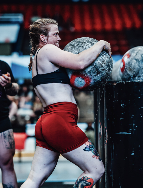 A woman lifting an atlas stone during a strongman competition in a gym.