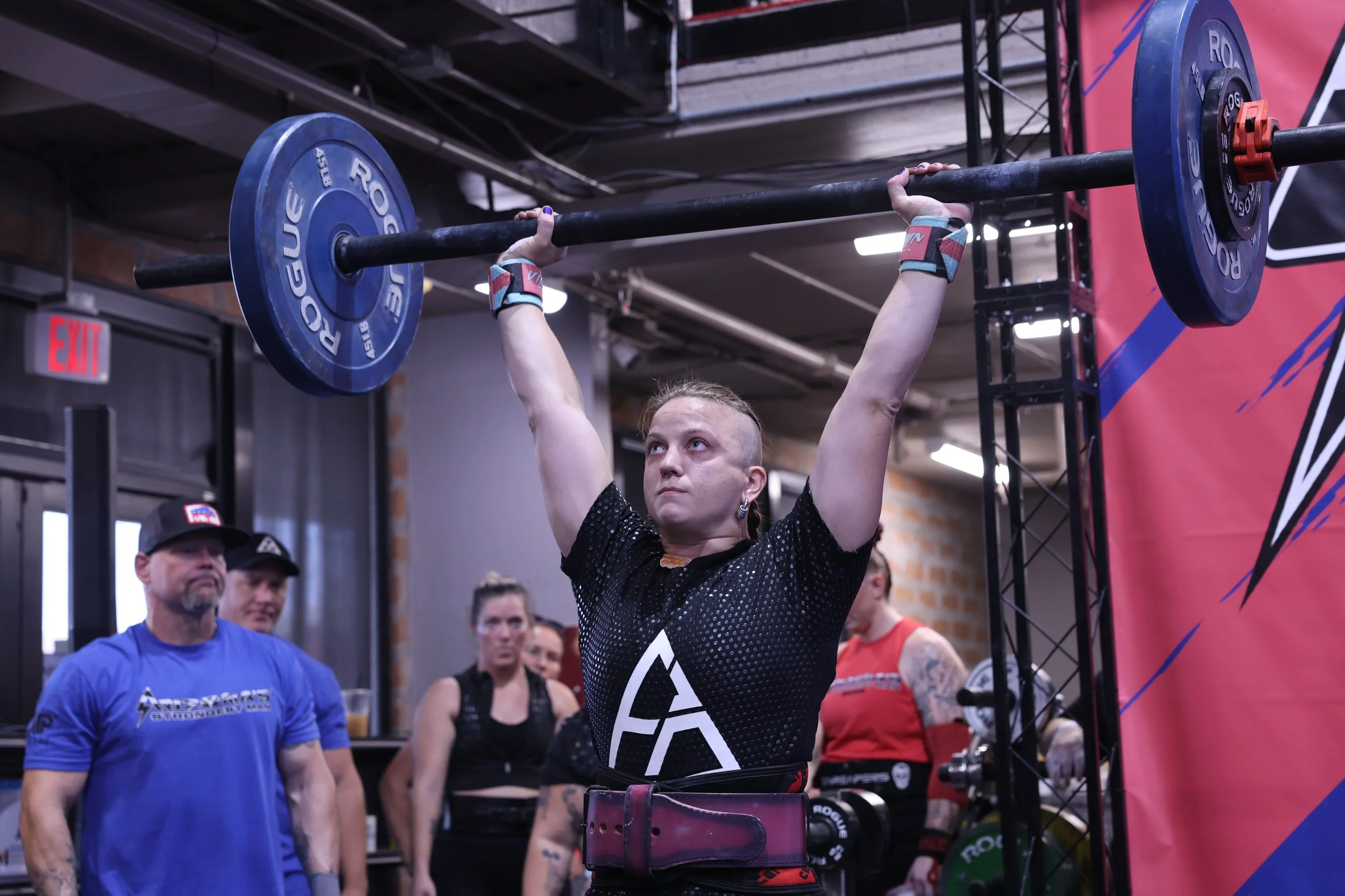 A woman lifting a barbell at a Strongman gym, surrounded by spectators and other participants.