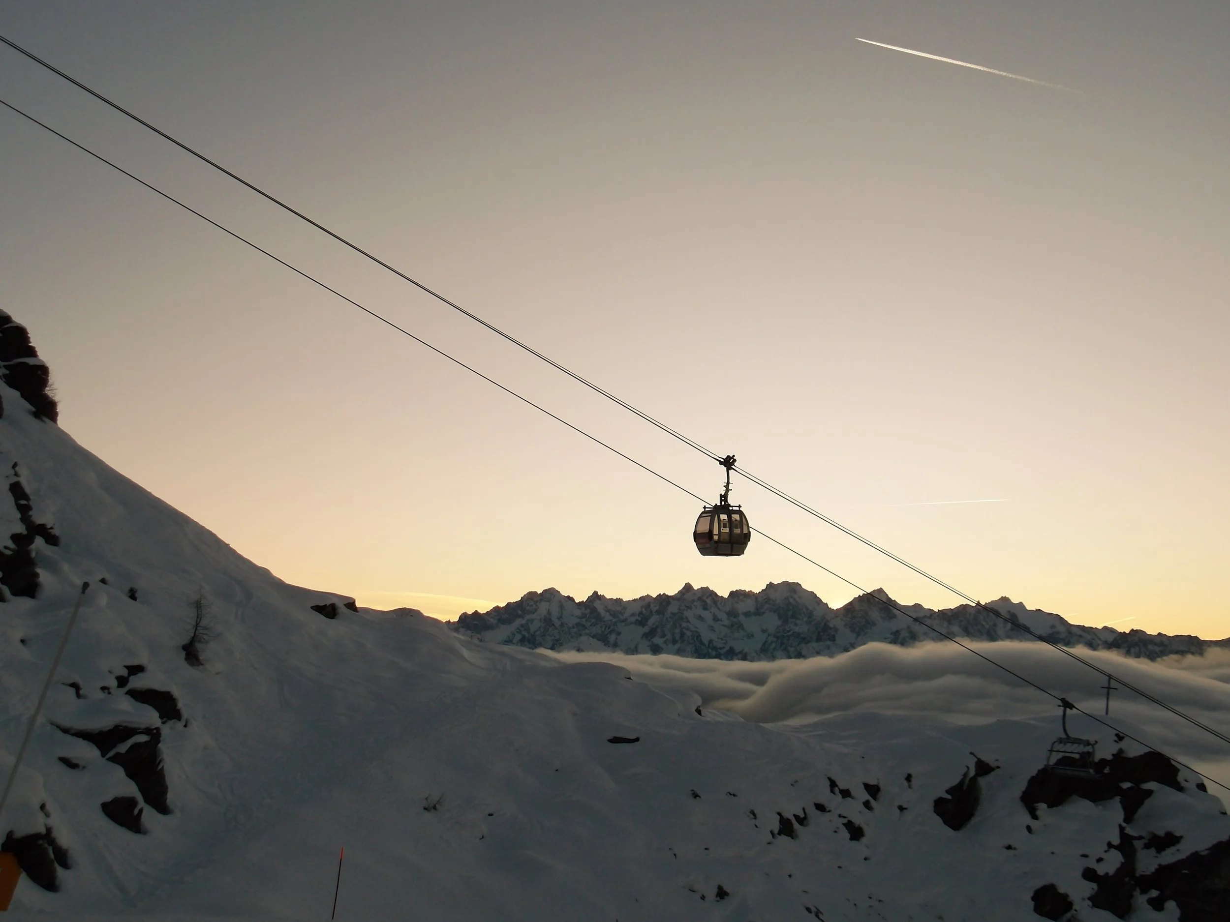 Snow-covered mountain landscape with a ski lift cable and gondola, set against a mountain range and a sky at dusk.