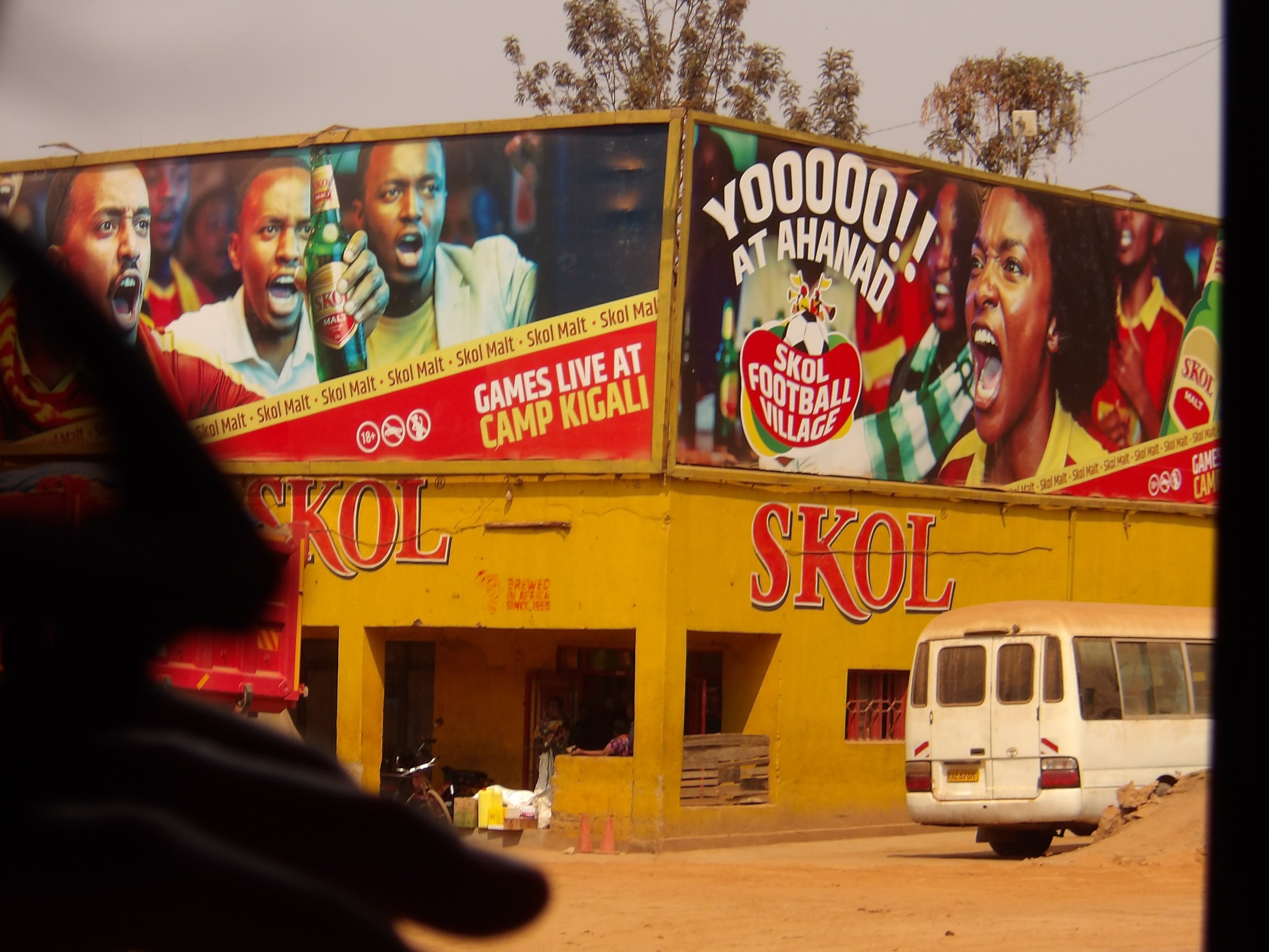 Billboard advertising SKOL beer with images of excited football fans and the text "YOOO! AT AHAND" and "GAMES LIVE AT CAMP KIGALI." The billboard is above a yellow building, and there are vehicles and people below.