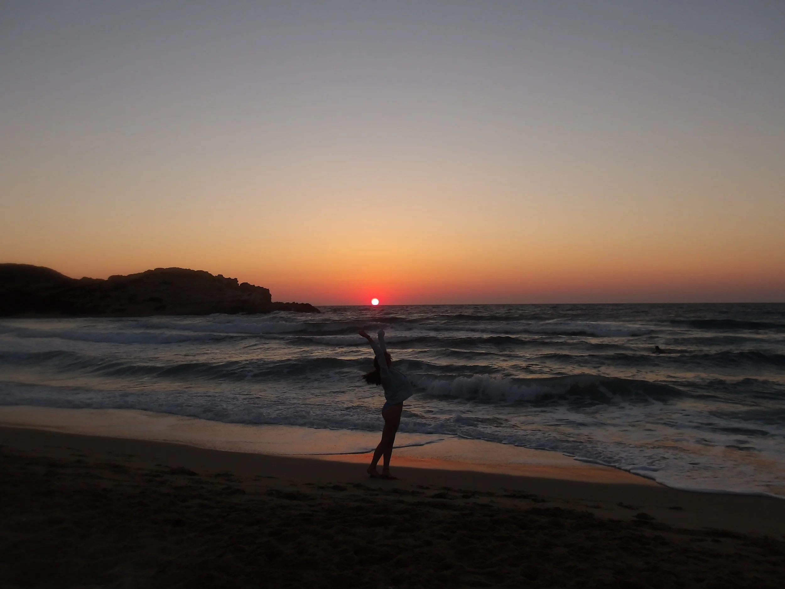 Person standing on the beach during sunset, with the sun near the horizon, and ocean waves in the foreground.