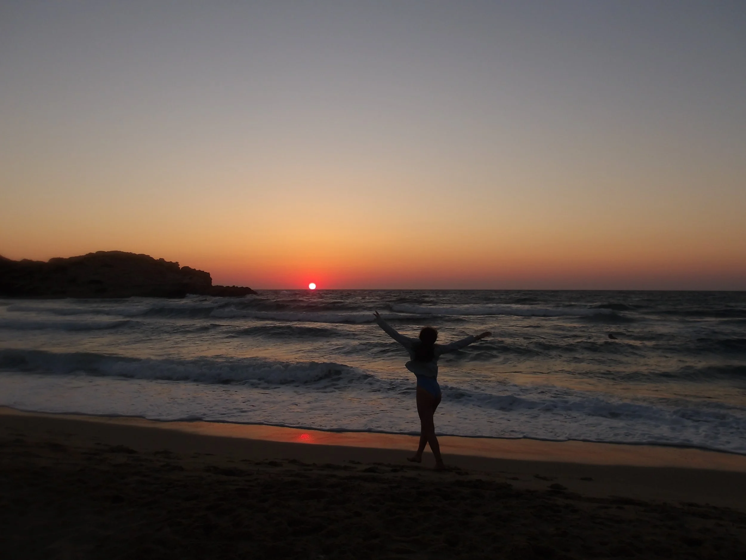 A child playing on the beach at sunset with arms outstretched, the sky transitioning from orange near the horizon to dark gray at the top, and the ocean waves in the background.