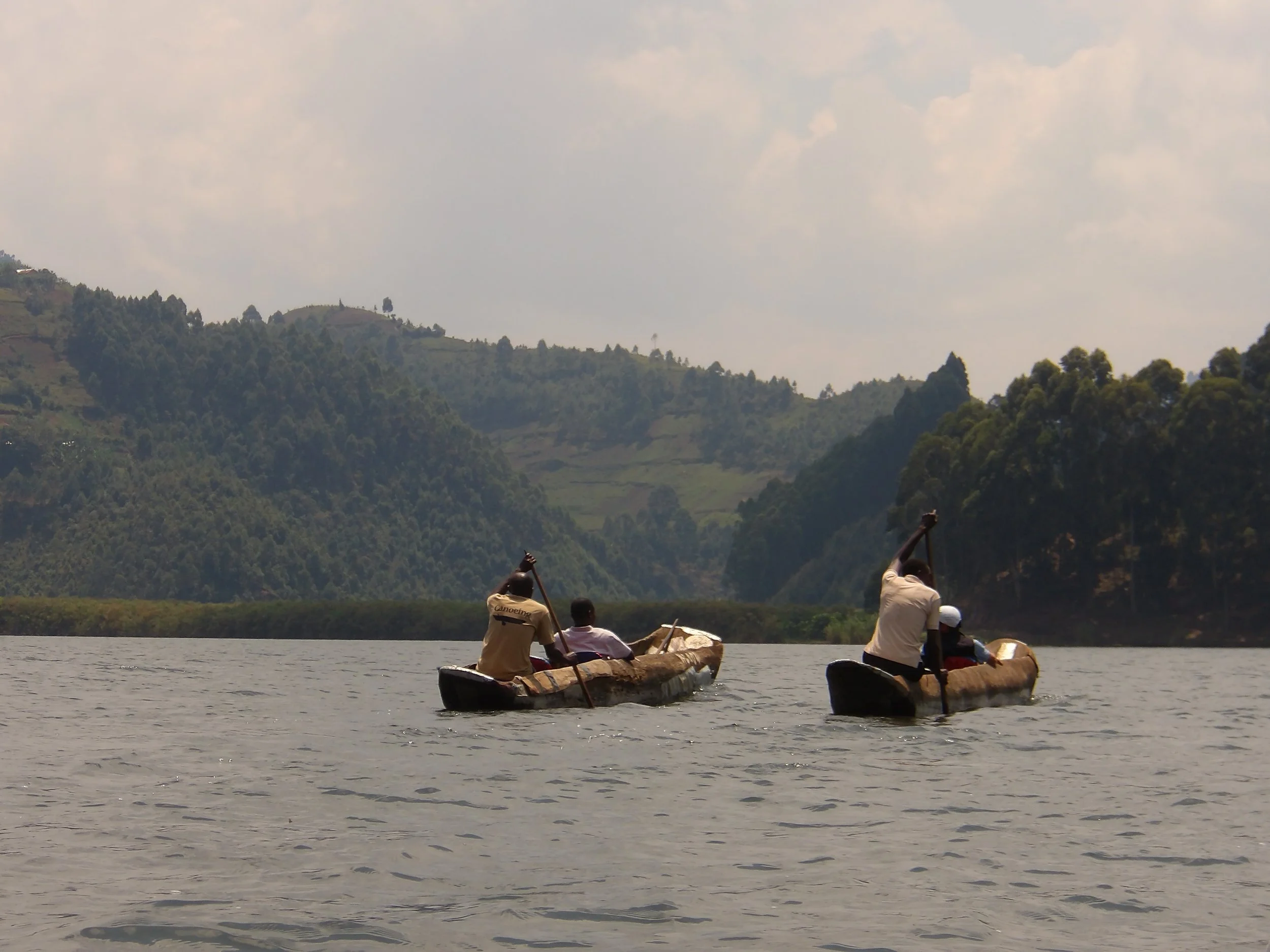Three people paddling traditional canoes on a river with green, hilly landscape and sparse trees in the background.