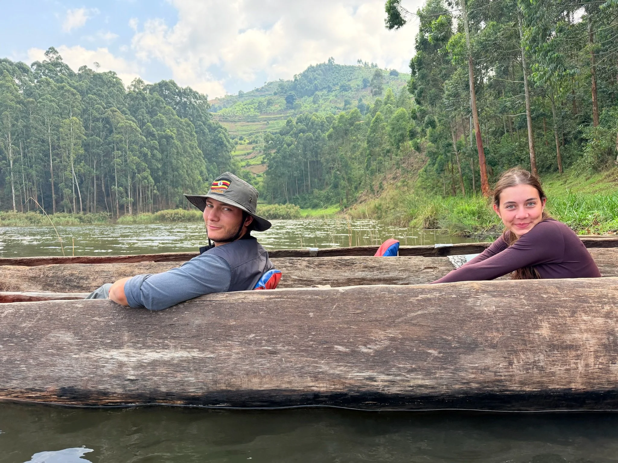 Two people sitting on a wooden boat on a river with lush green forested hills in the background, one man wearing a wide-brimmed hat and a woman with brown hair smiling.