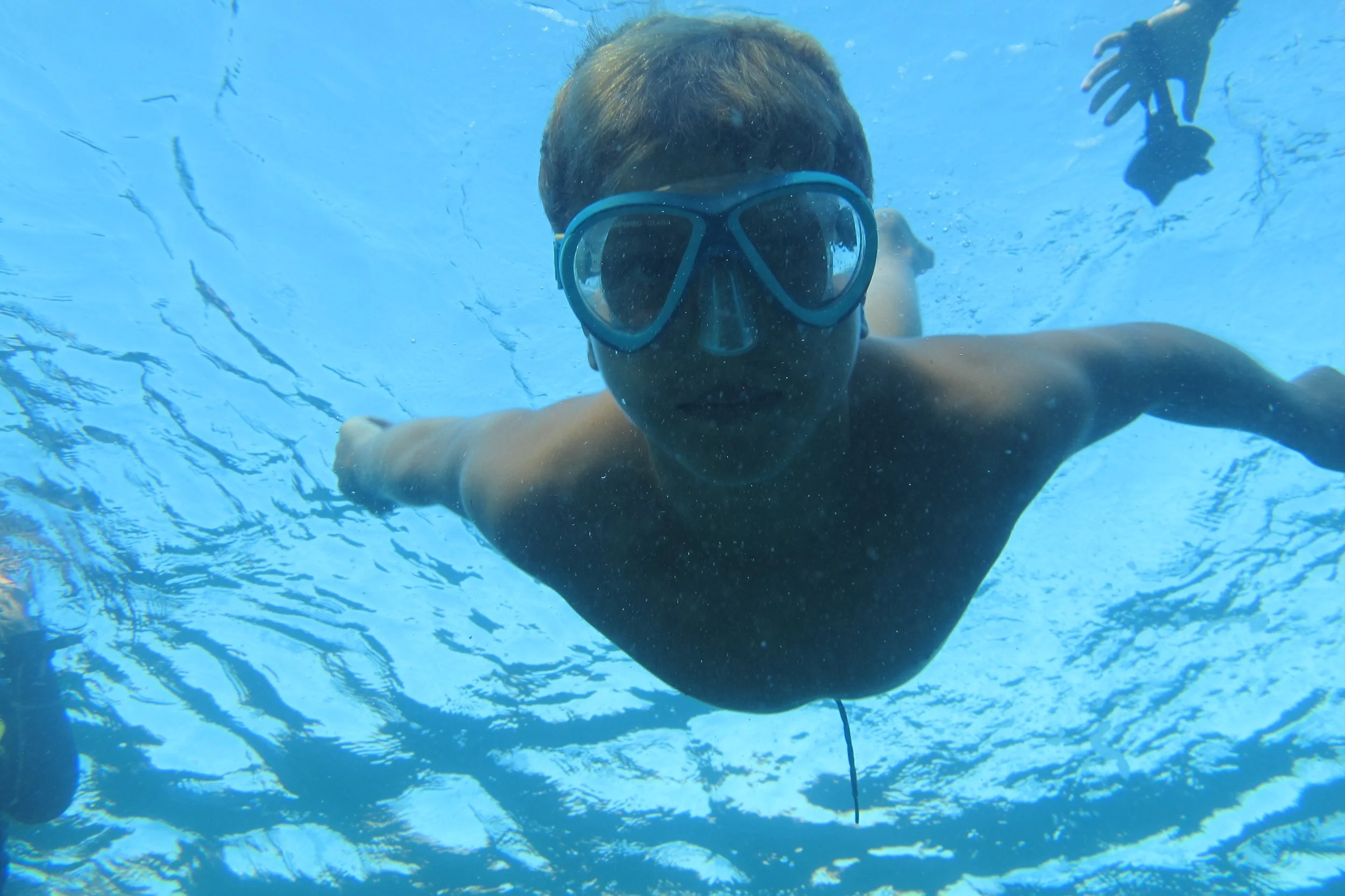 Person swimming underwater, wearing a swim mask and reaching out with arms extended, with blue water and ripples around.