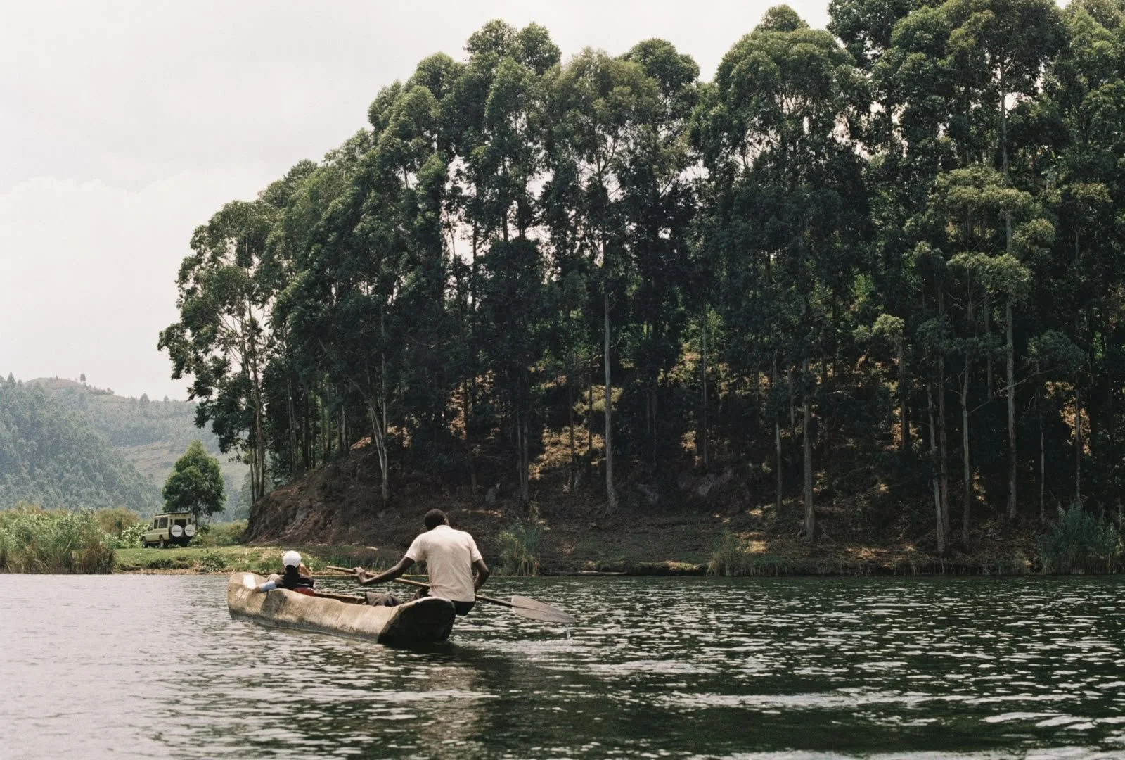 Two people are in a canoe on a river, with a man sitting at the front and a man at the back rowing, surrounded by tall green trees on the riverbank.