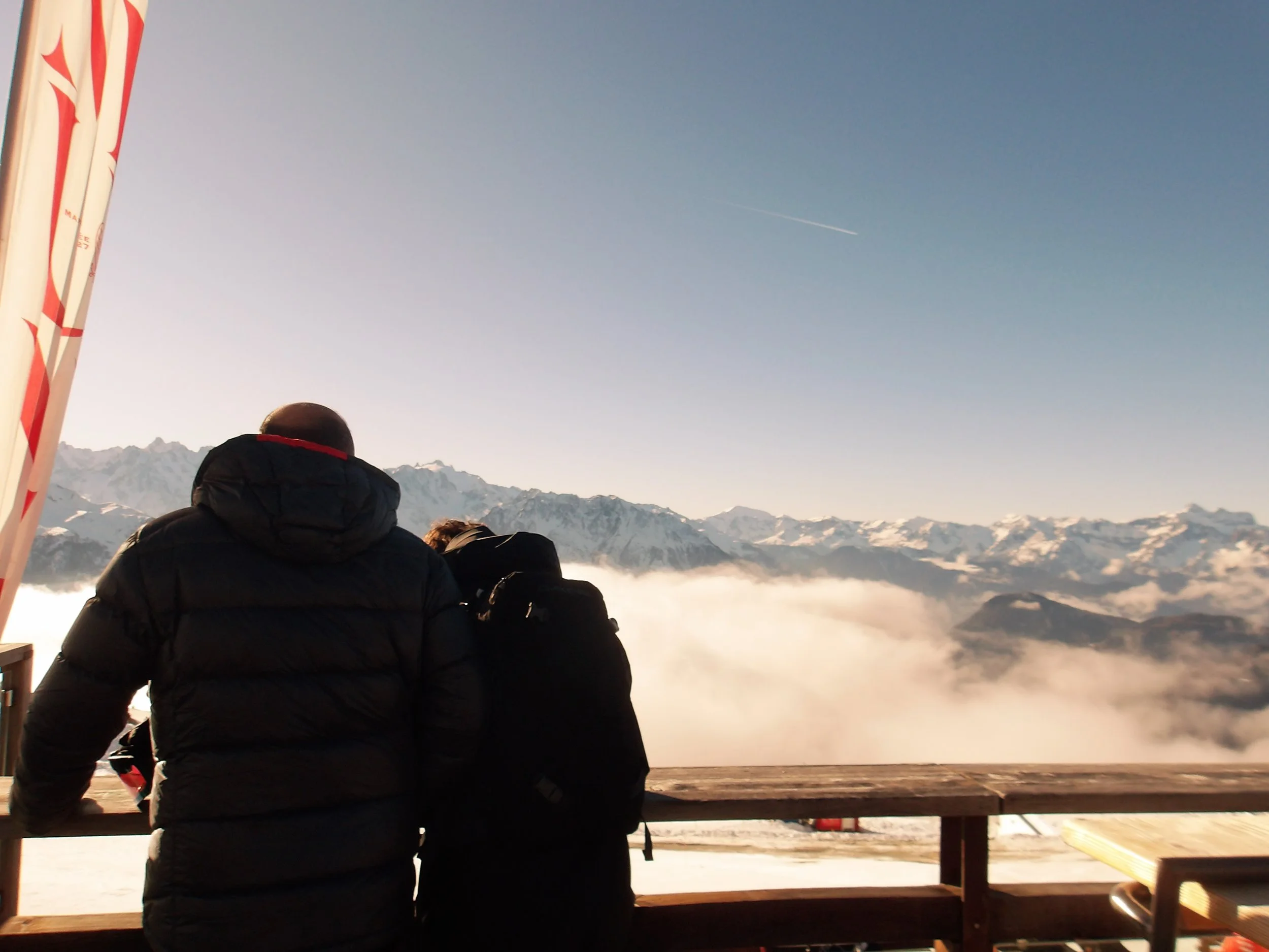 People sitting on a wooden deck overlooking a mountain range with snow-capped peaks, dressed in winter clothing, enjoying the view of the sky and a jet trail.