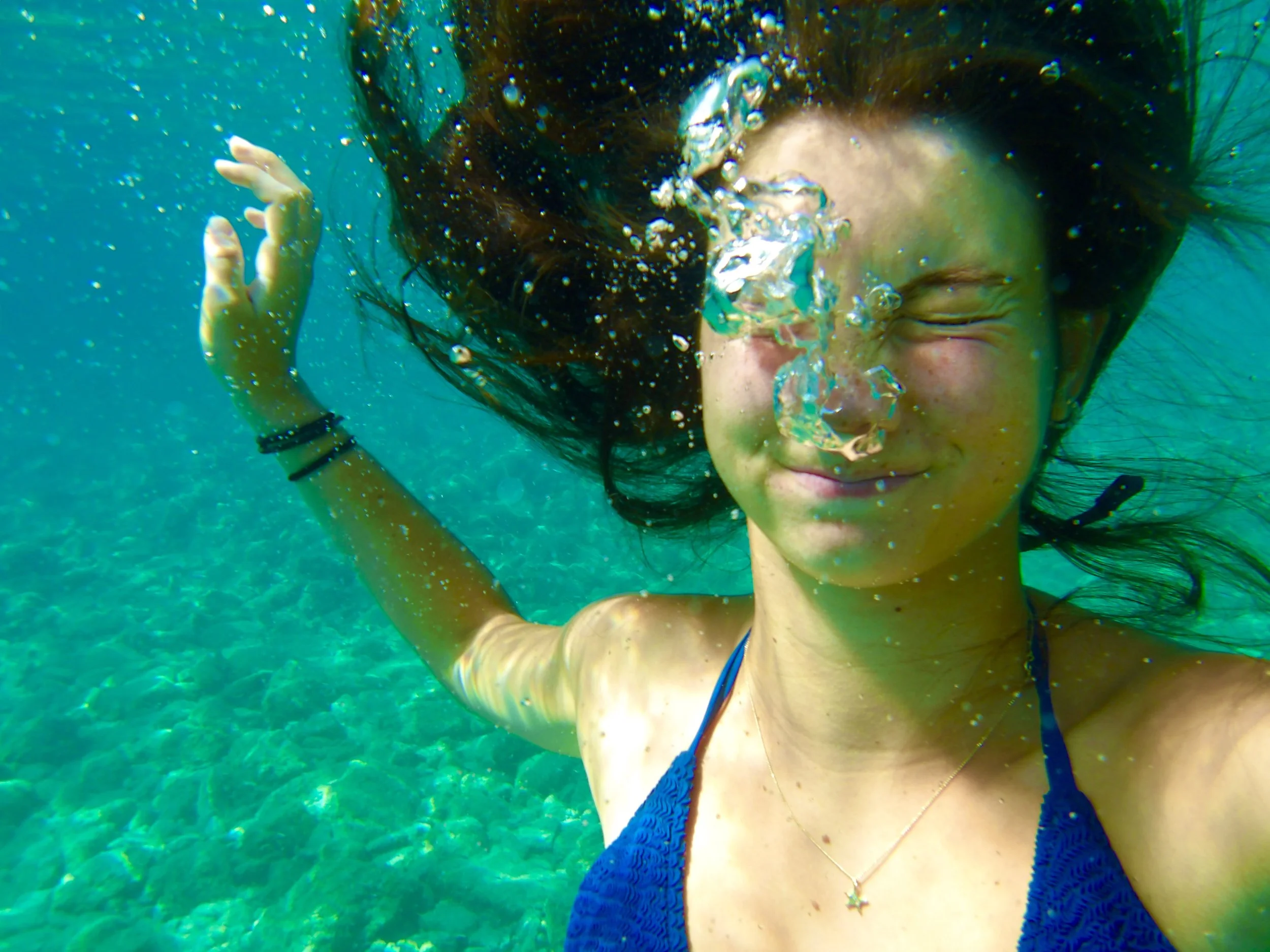 A woman with dark hair swimming underwater, wearing a blue bikini, with her eyes closed and a smile on her face, surrounded by bubbles.