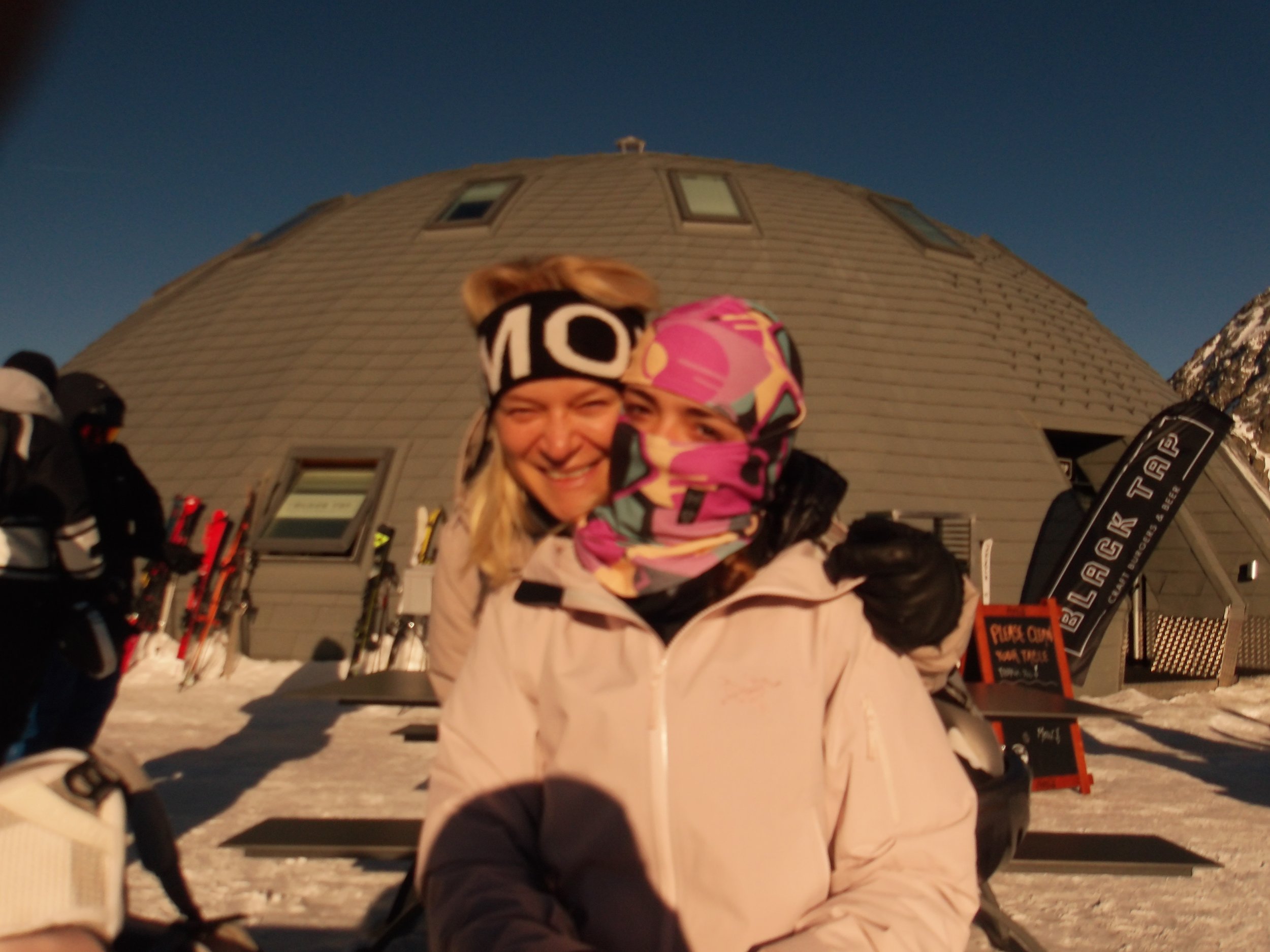 Two people smiling, dressed in winter gear, with a domed building in the background at sunset or early evening.