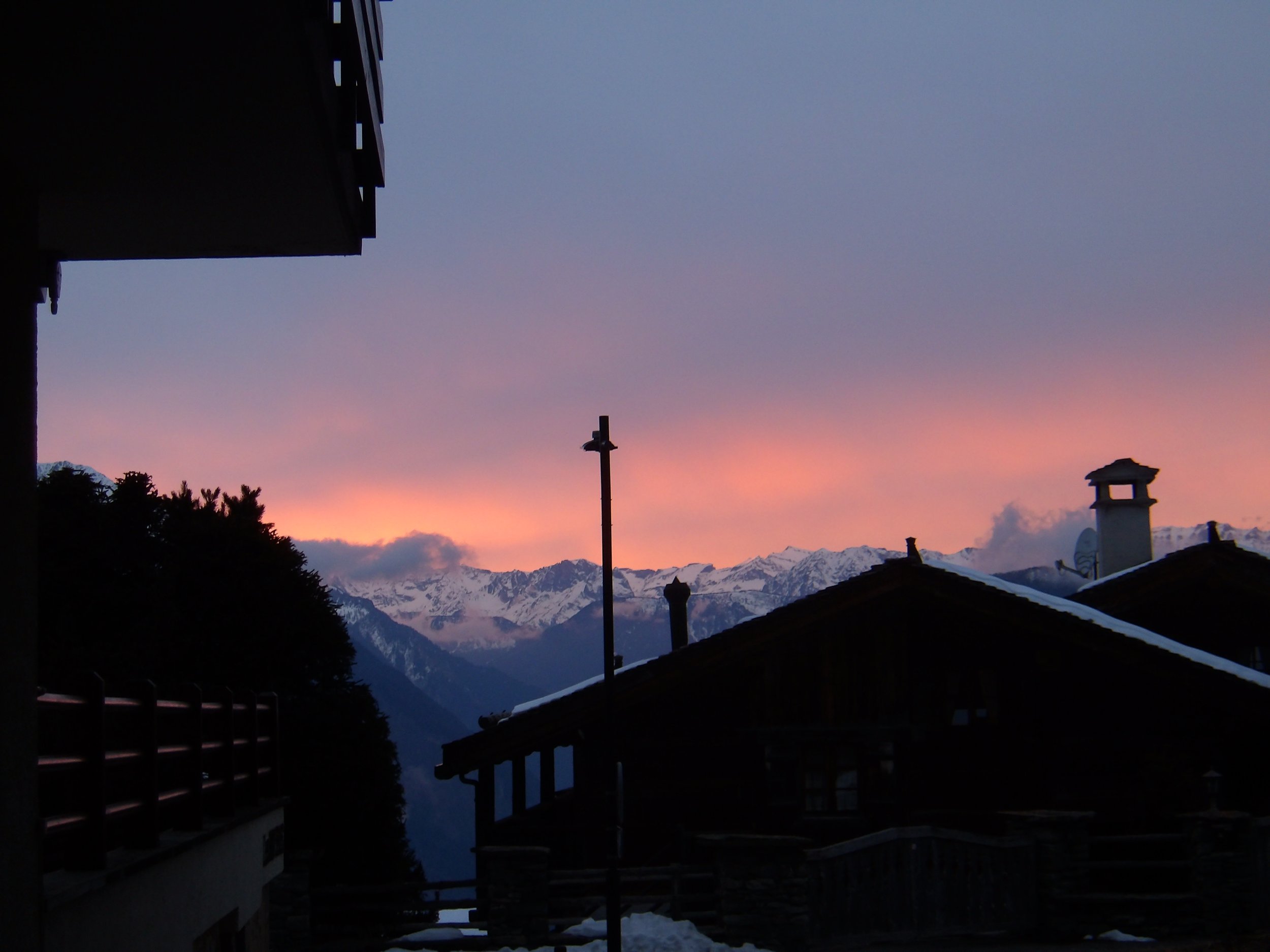 Scenic view of snow-capped mountains during sunset, with rooftops and silhouettes of trees and buildings in the foreground.