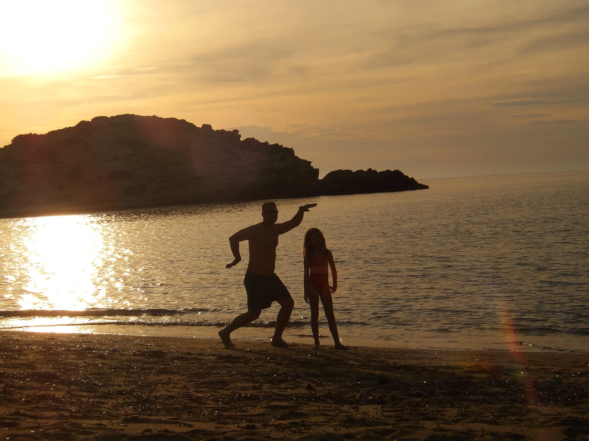 A man and a woman are on the beach during sunset, with a large hill or small mountain in the background. The man is shirtless and appears to be dancing or running, while the woman stands nearby with her back to the camera. The sunlight reflects off the water, creating a warm glow.
