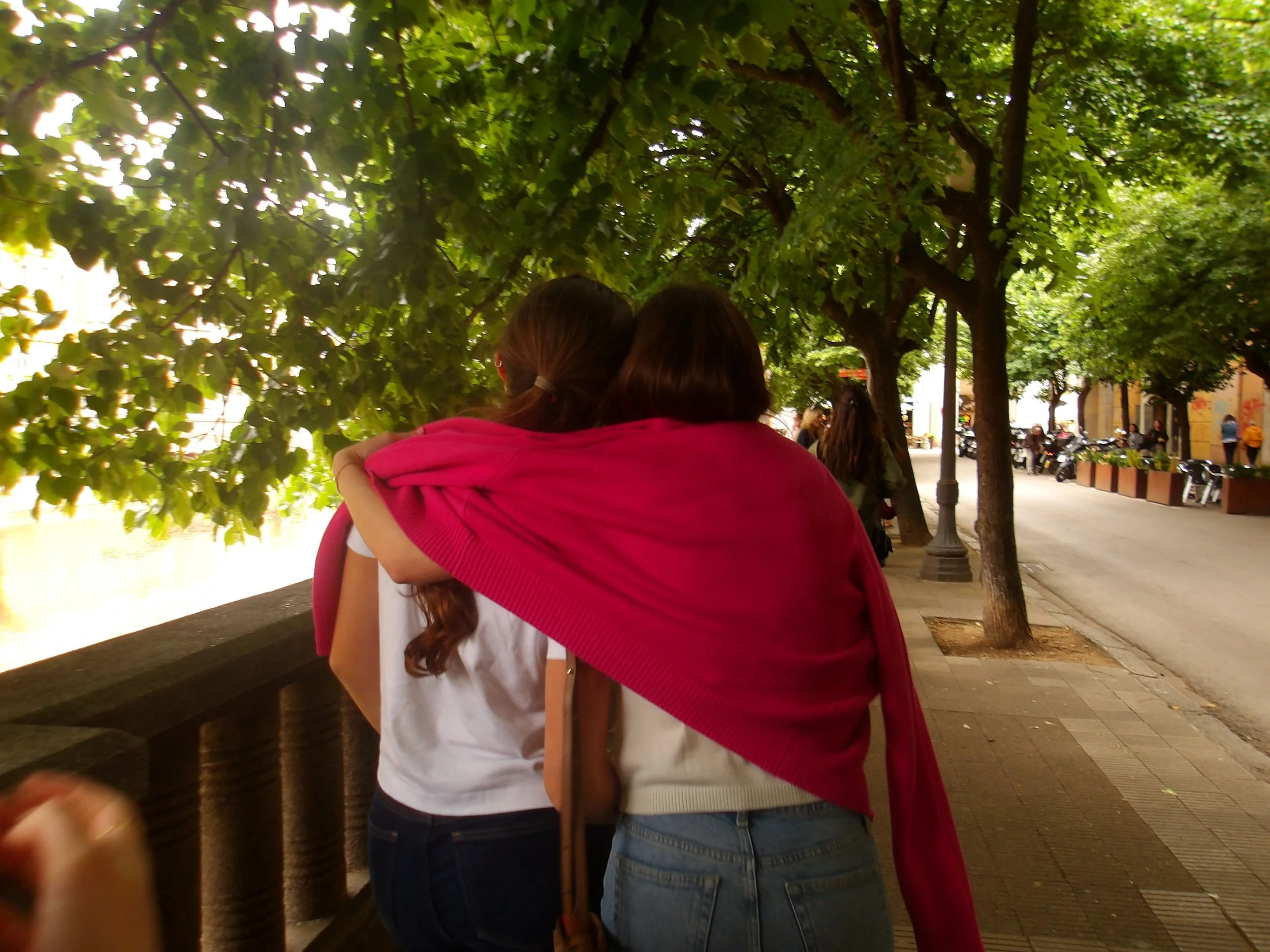 Two women embrace on a sidewalk near trees with green leaves, with motorcycles and pedestrians in the background.