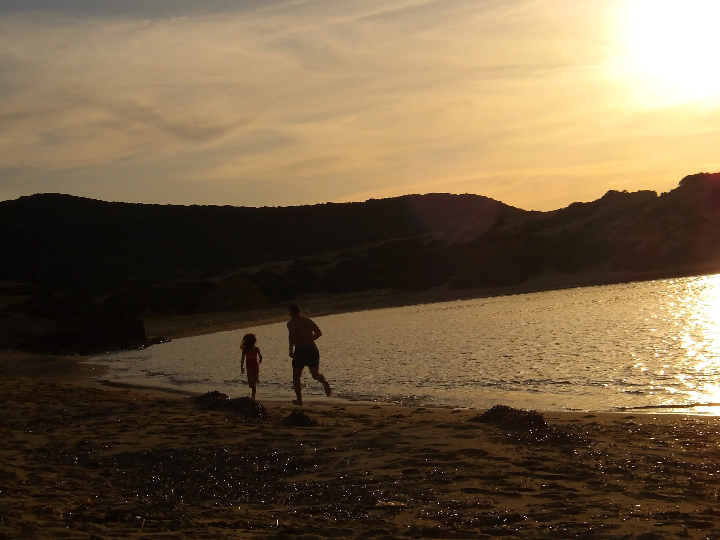 A silhouette of a person and a child walking along the beach at sunset, with hills and a partly cloudy sky in the background.