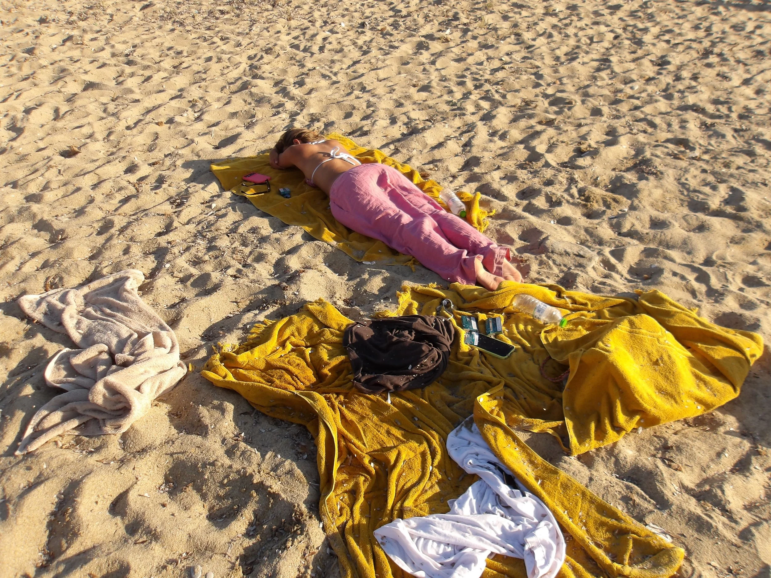 A person is lying face down on a yellow towel on sandy beach, surrounded by scattered clothing and water bottles.