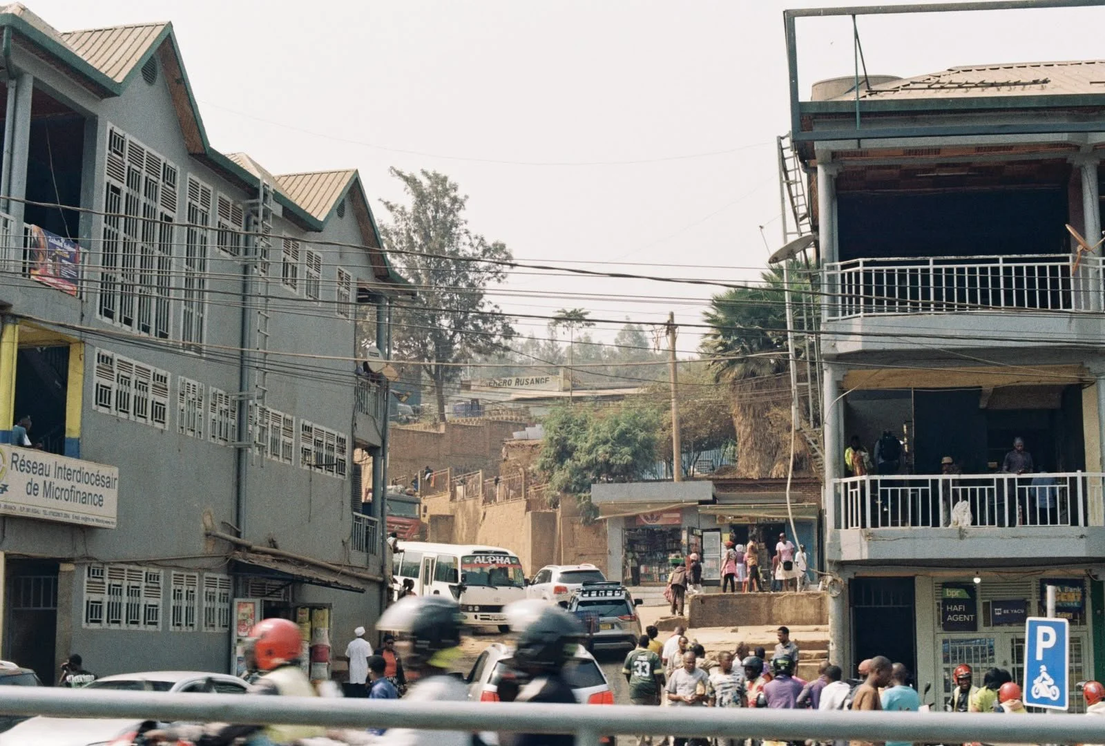 Busy street scene with cars, motorcycles, and pedestrians in an urban area with multi-story buildings and trees in the background.