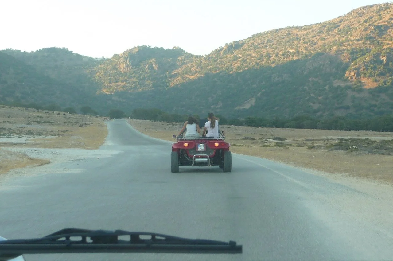 Two women riding on a red ATV on a winding rural road with mountains in the background.