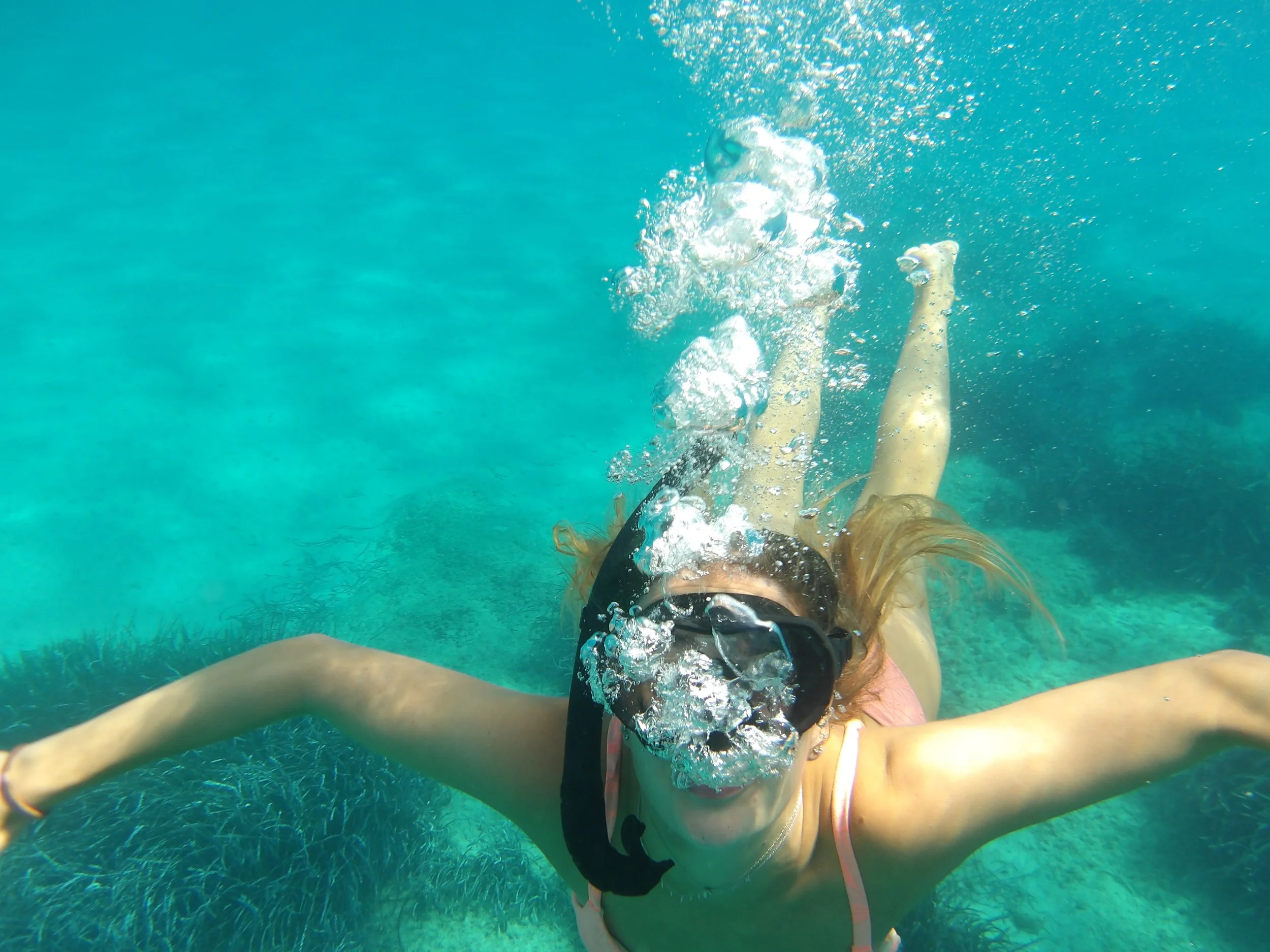 A person swimming underwater, reaching out with their arms. The person is wearing a swimsuit and a mask, with bubbles around their head and arms. The background features clear, turquoise water and underwater vegetation.