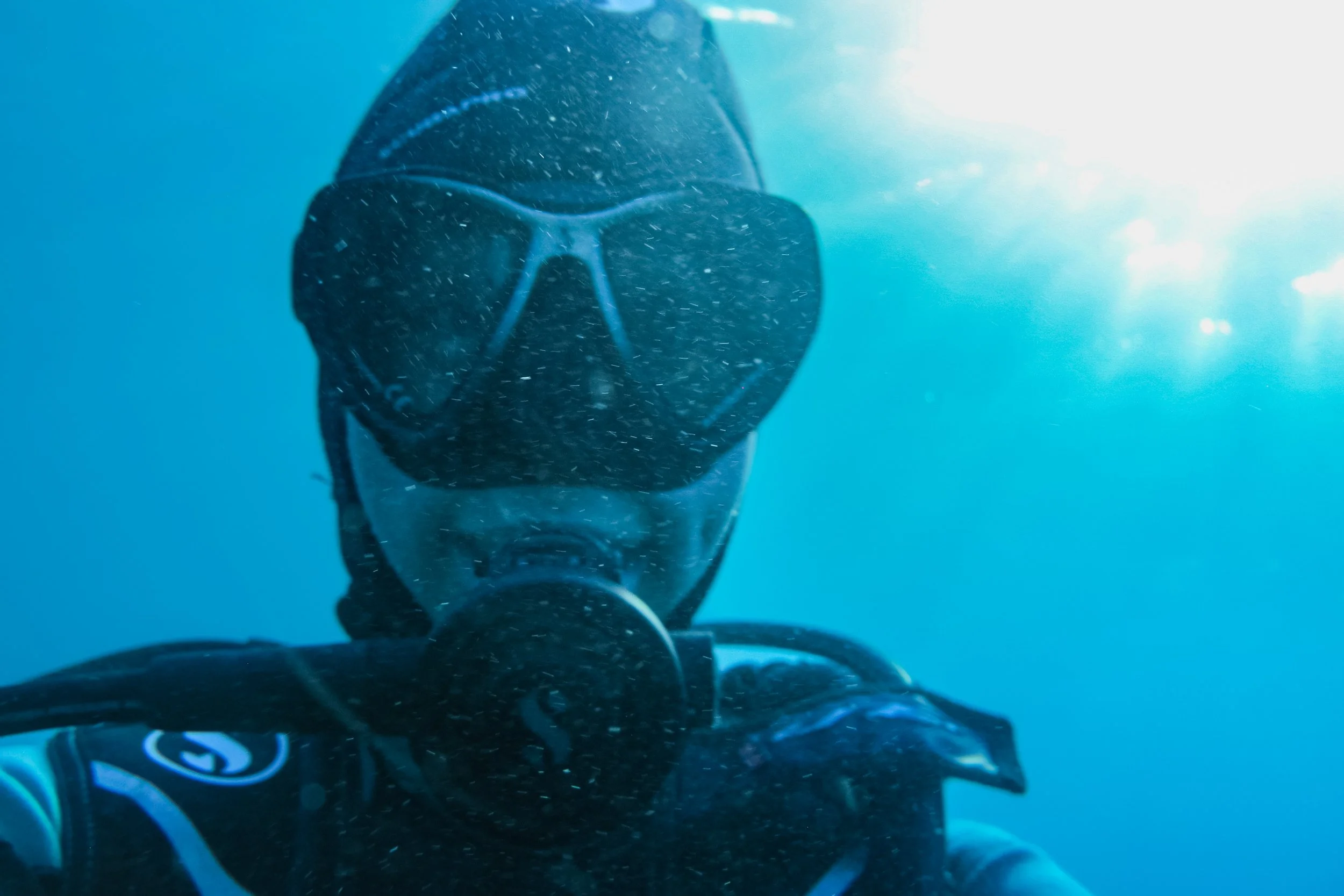 A person in scuba gear underwater, wearing a diving mask and a wetsuit, with sunlight piercing through the water.
