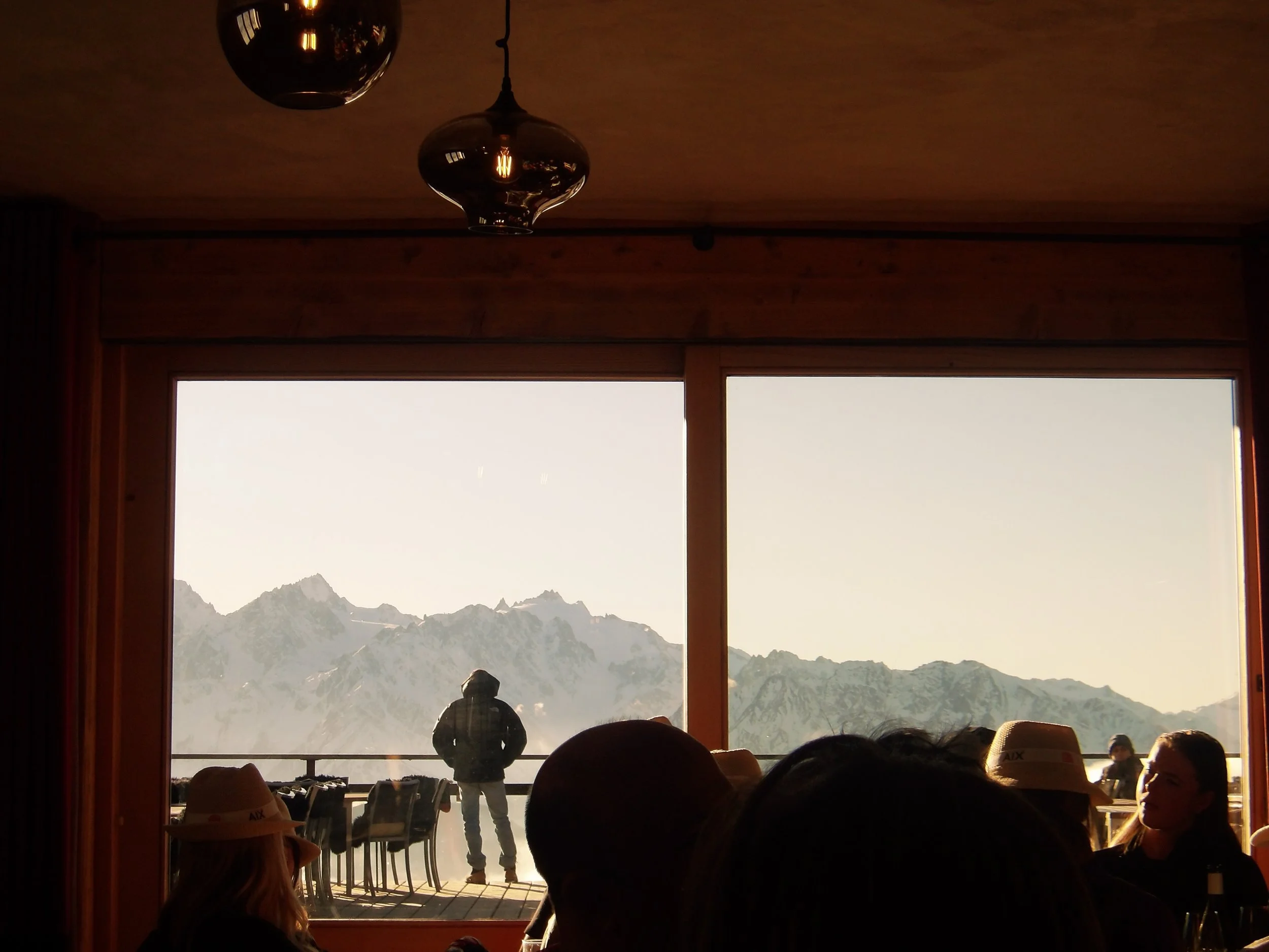 People sitting indoors near a large window overlooking snow-capped mountains and a person standing outside in front of the window, with a mountain range in the background.
