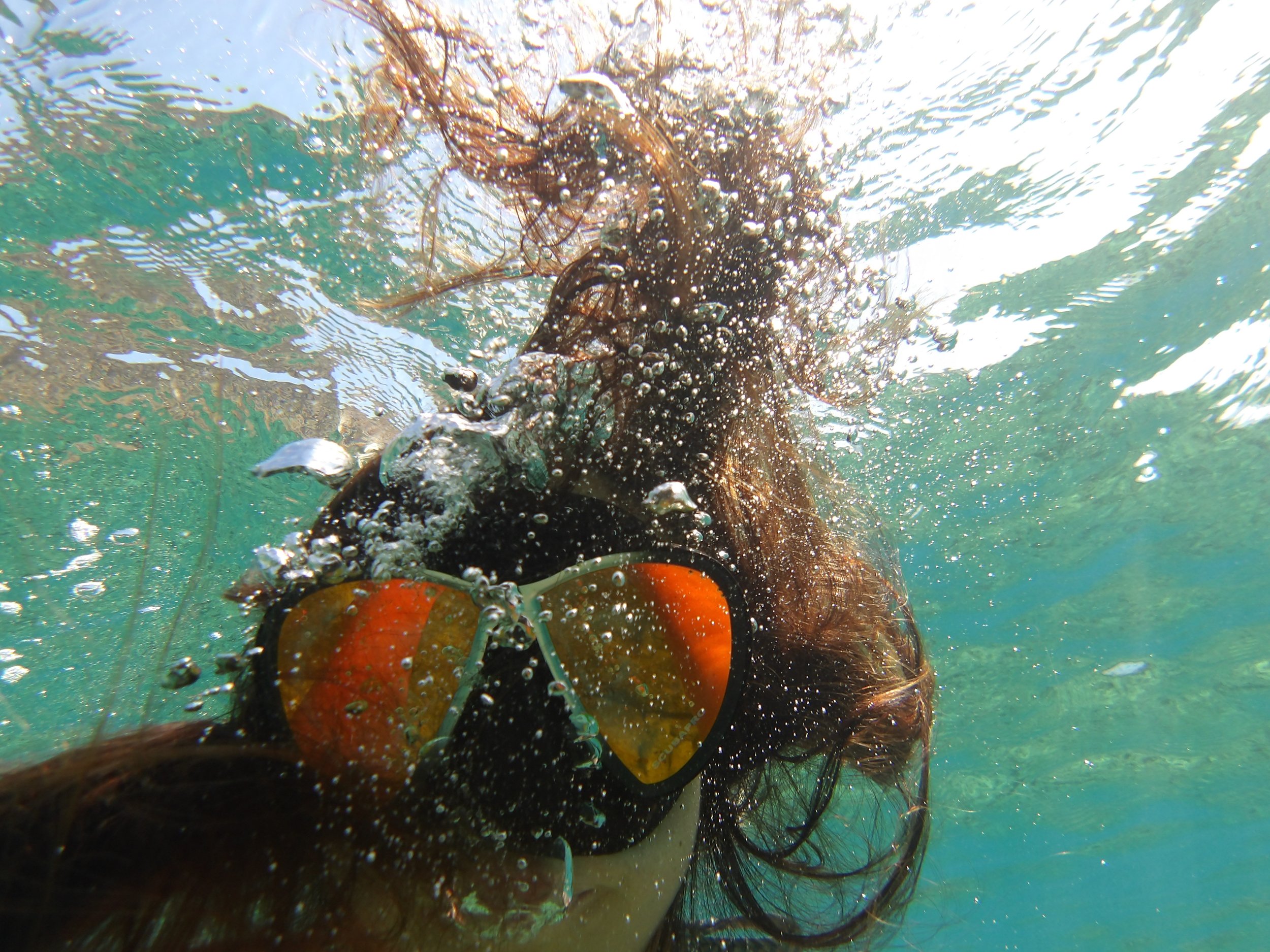 A person with long hair swimming underwater, holding onto a bright orange buoy with a yellow and black strap. The scene captures bubbles and water movement in a greenish-blue aquatic environment.