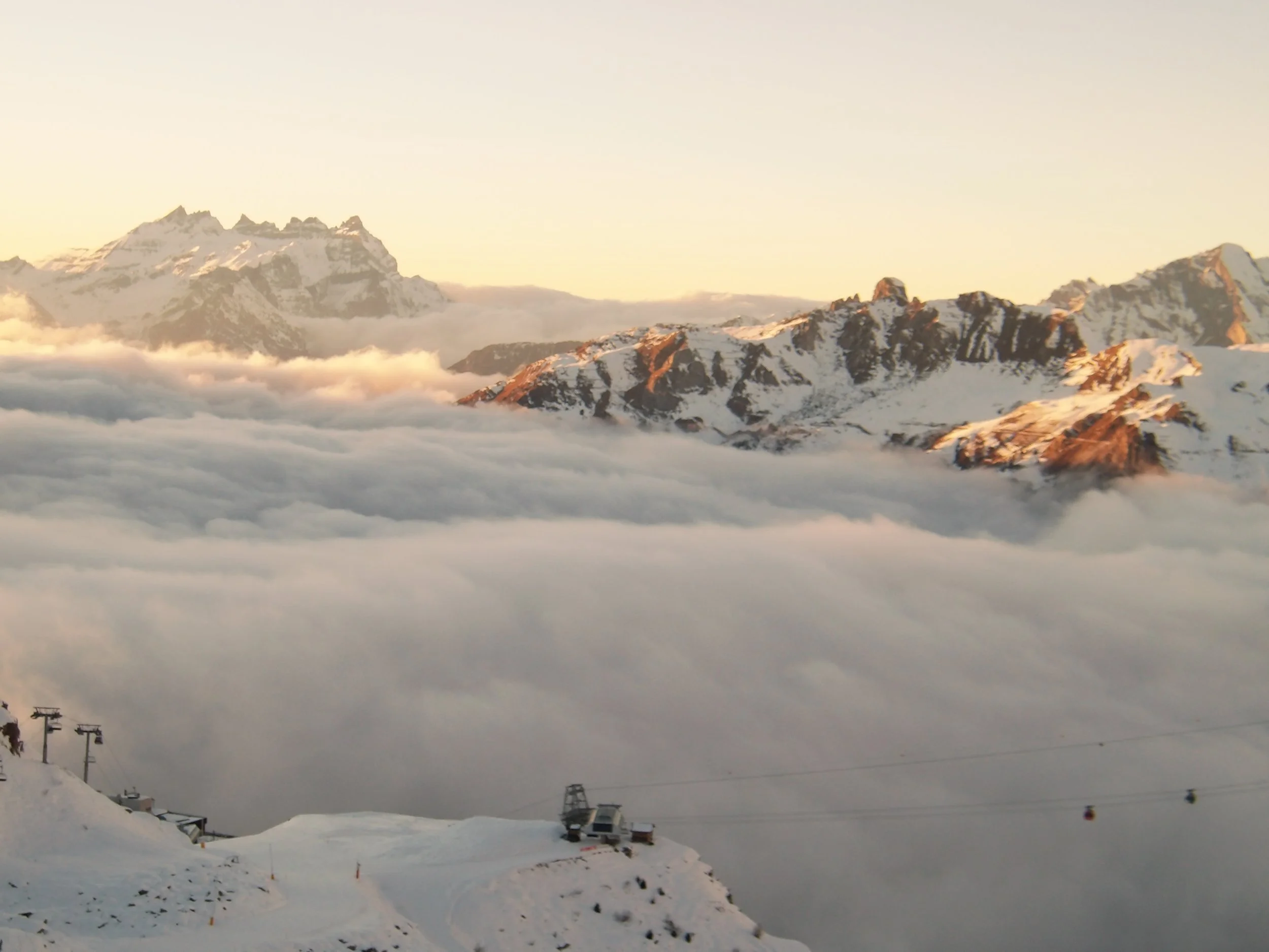 Mountain snow peaks with clouds around them, ski lifts in the lower snow-covered area, and a ski resort at the base.