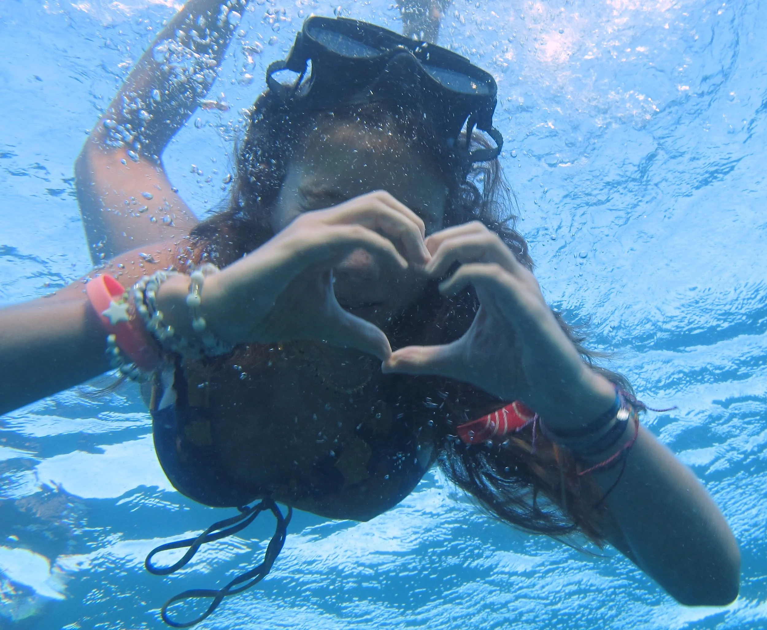 Person underwater wearing snorkeling gear, making a heart shape with their hands, surrounded by water bubbles.