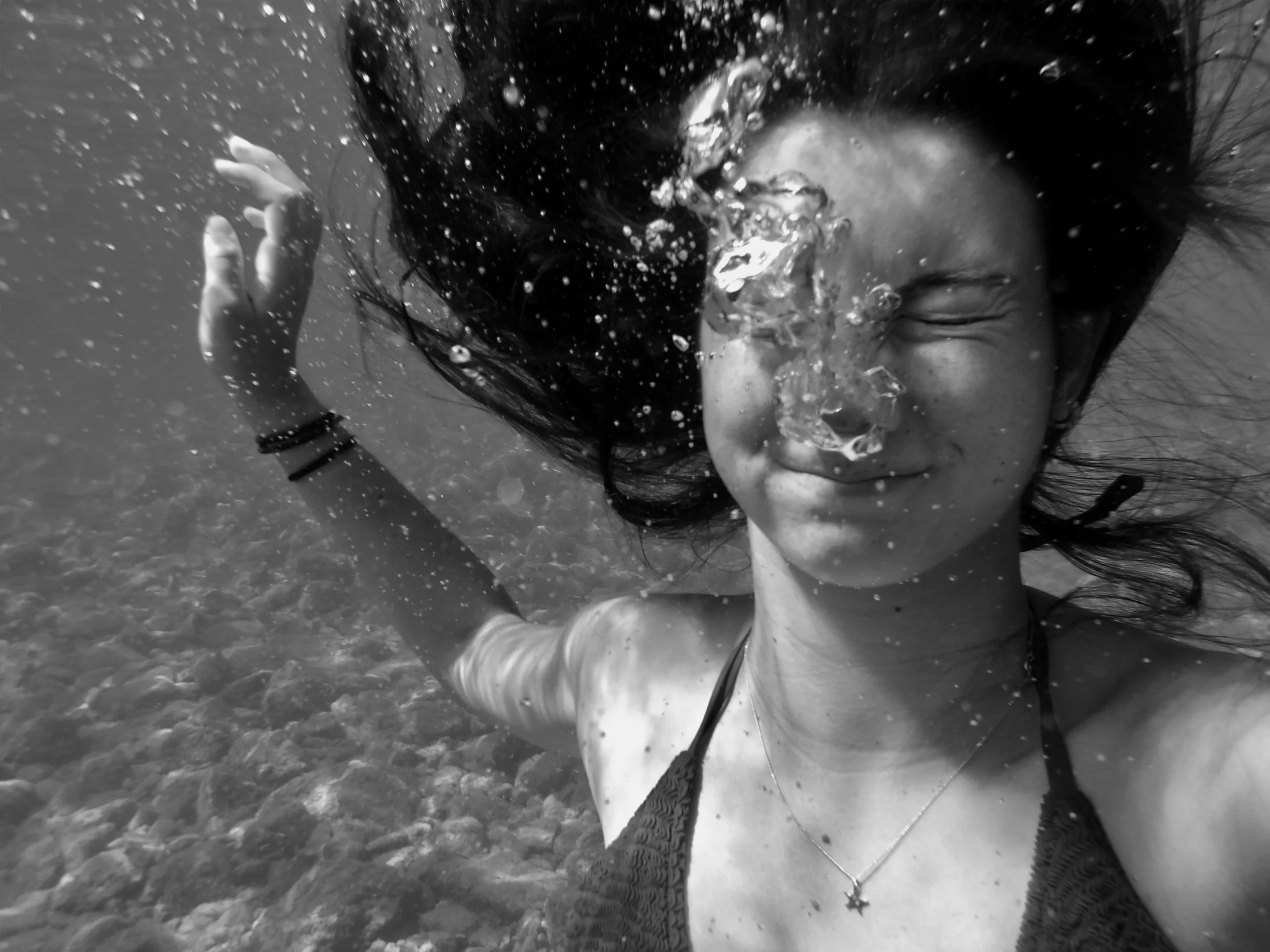 A woman swimming underwater, smiling with her eyes closed, with dark hair flowing around her face, surrounded by bubbles, in a black-and-white photo.