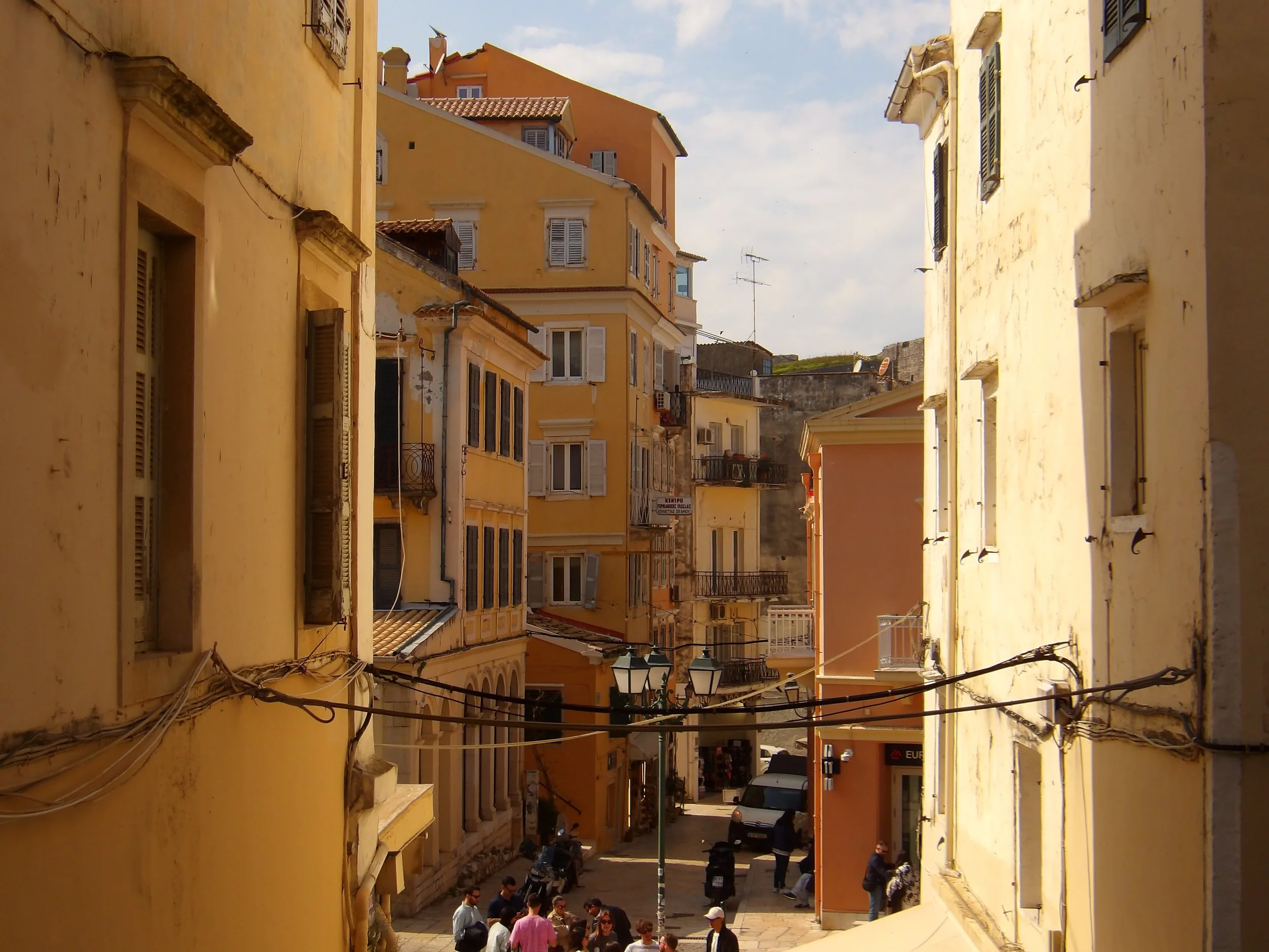 A narrow street scene in a European town with yellow and orange buildings, some with window shutters, and shops at the ground level. There are people walking and socializing on the street, and cars parked along the street. The sky is partly cloudy.