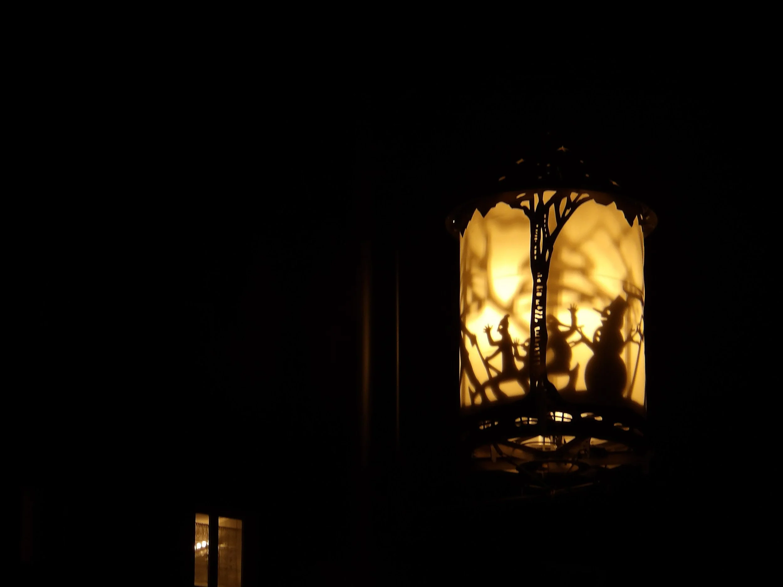 A decorative lantern with intricate cutouts is lit from within, casting shadows of trees or branches on its surface. The lantern emits a warm, yellow glow against a dark background.