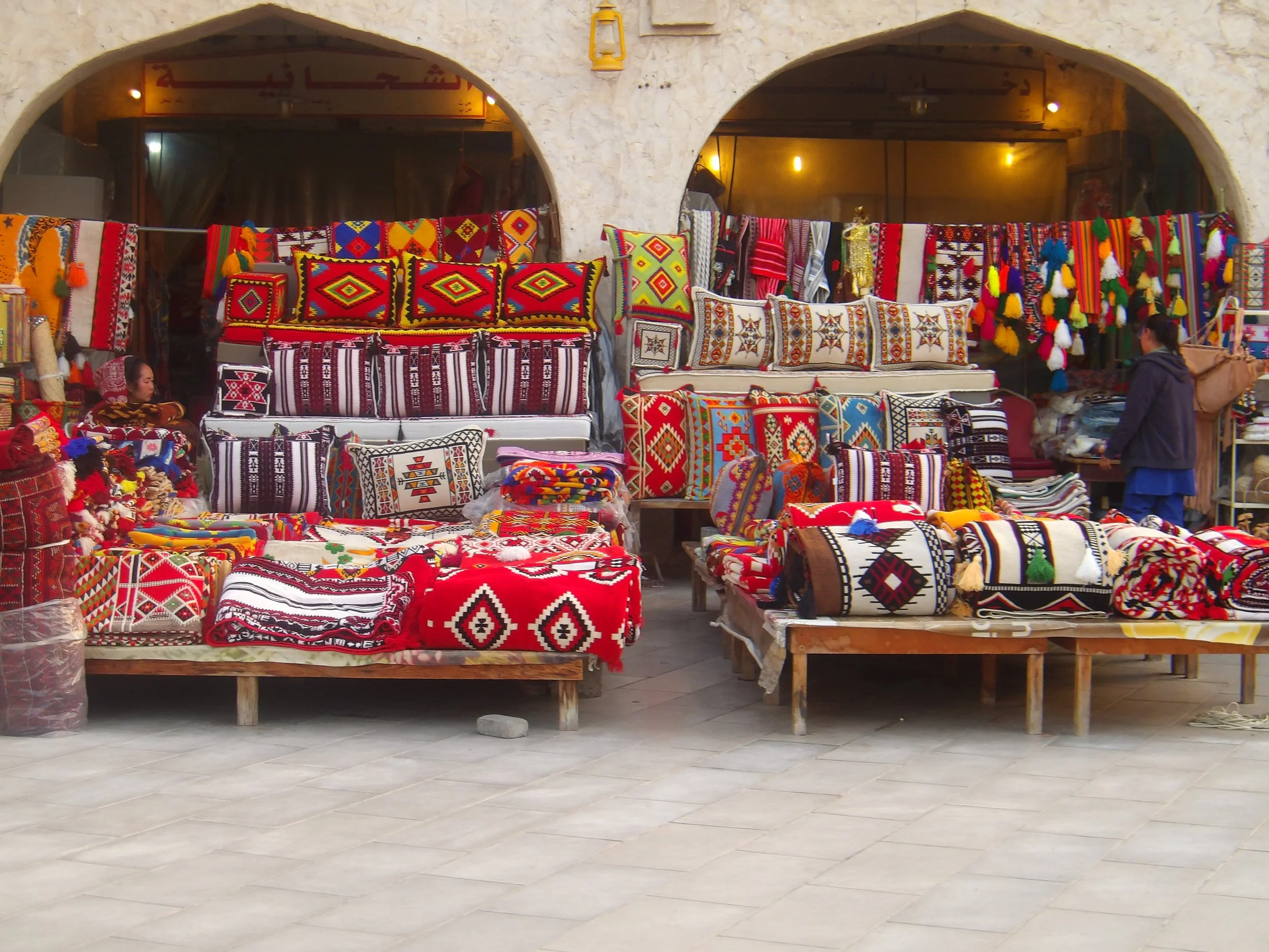 Market stall selling colorful traditional woven textiles, pillows, and bags in an outdoor setting with arches in the background.