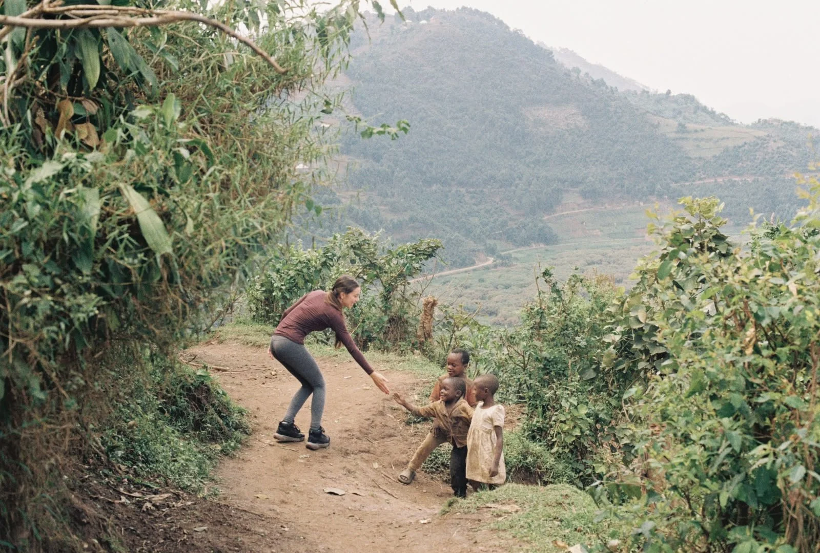 A woman playing with three children on a dirt path in a rural, hilly landscape with lush green bushes and mountains in the background.