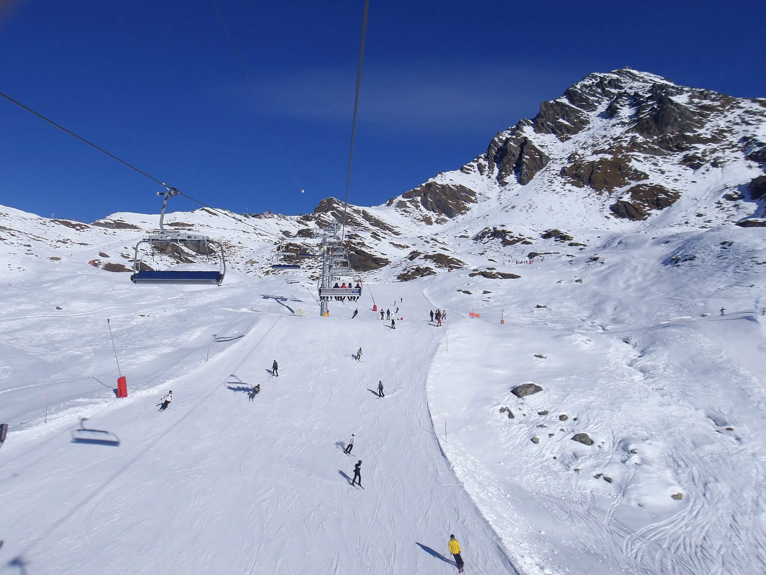 Snow-covered ski resort with chairlifts, skiers, and snowboarders on slopes, surrounded by mountainous terrain under a clear blue sky.