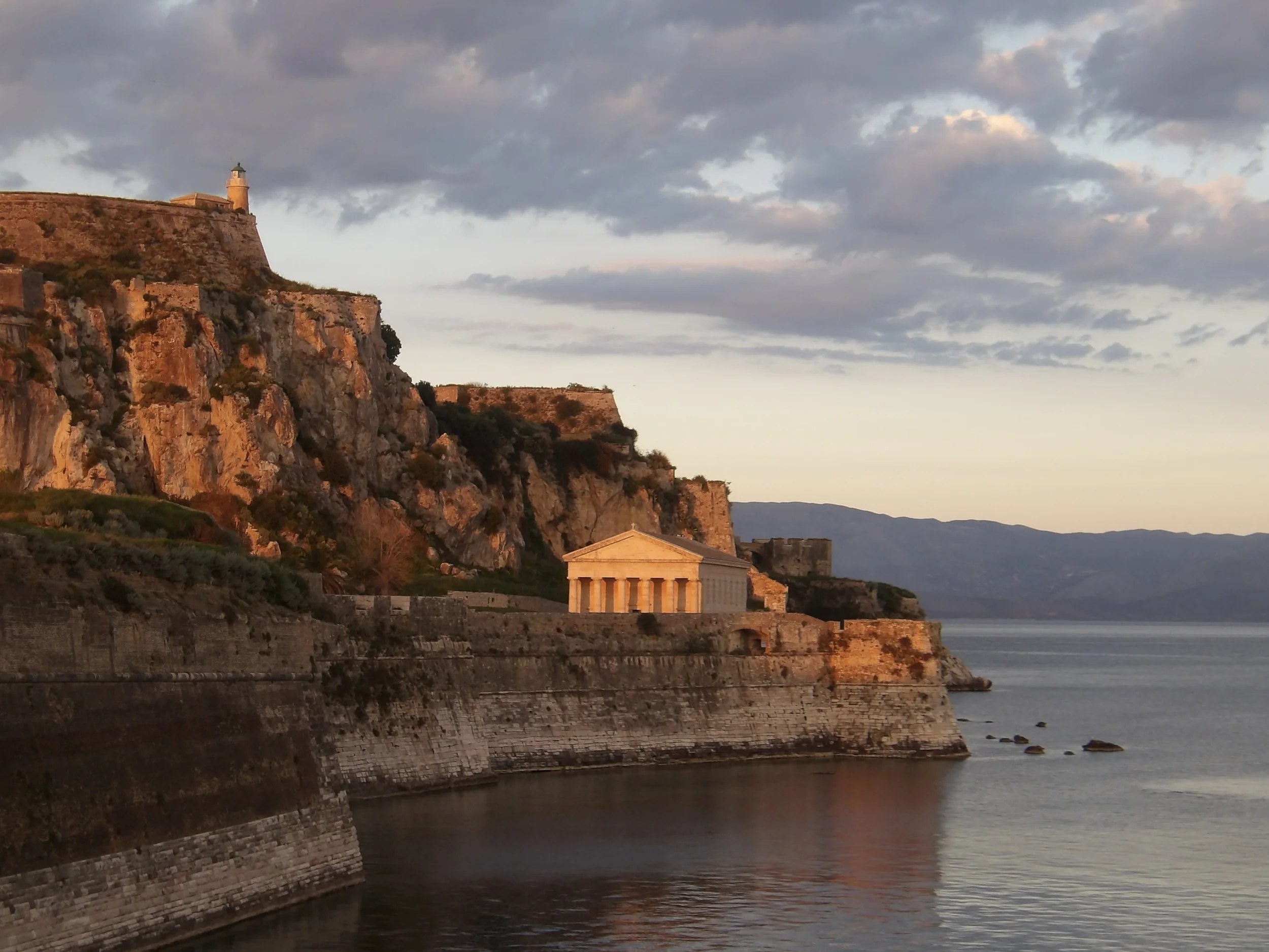 A coastal scene at sunset featuring a rocky cliff with a lighthouse on top, a classical building with columns near the water, and a calm body of water reflecting the sky and cliffs, with a cloudy sky overhead.