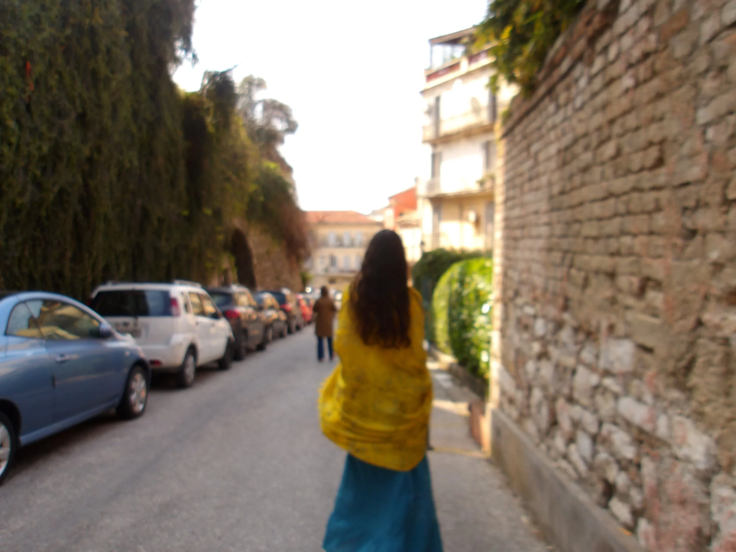 Person walking along a street with parked cars and buildings on a sunny day.