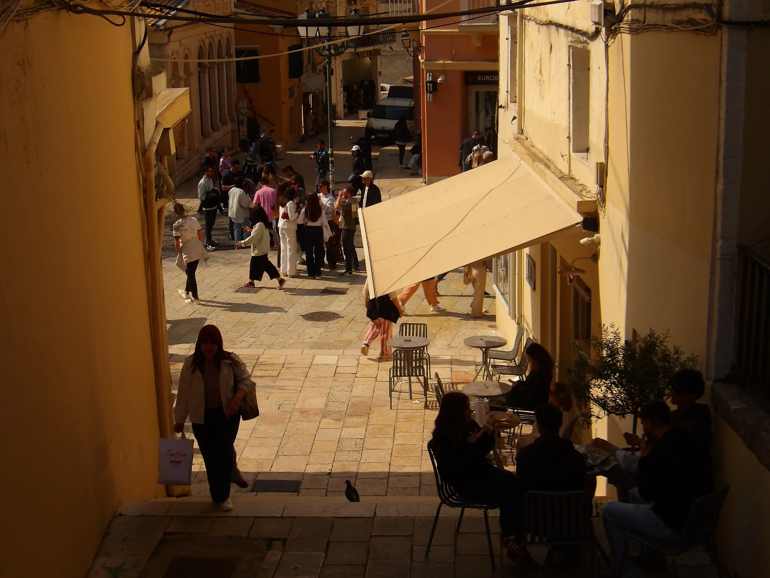 Outdoor street scene with people walking and sitting at tables, yellow buildings, and a small narrow street in a European city during daytime.