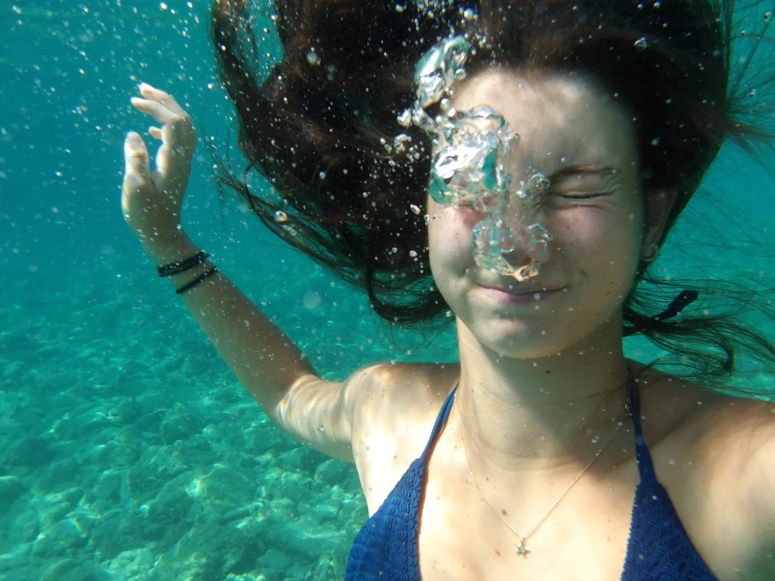 A woman underwater with her eyes closed, wearing a blue bikini and jewelry, surrounded by bubbles.