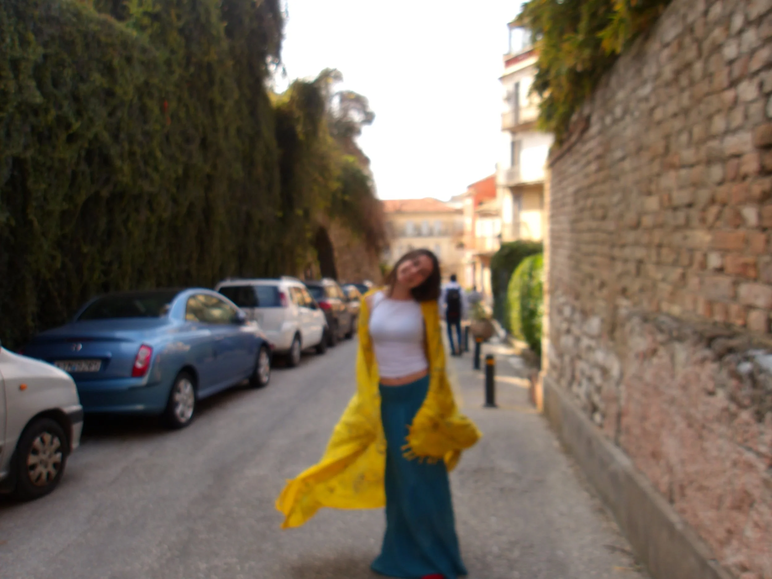 A young woman is walking on a sidewalk next to a stone wall with residential buildings in the background. Cars are parked along the street, and there are trees on the opposite side.