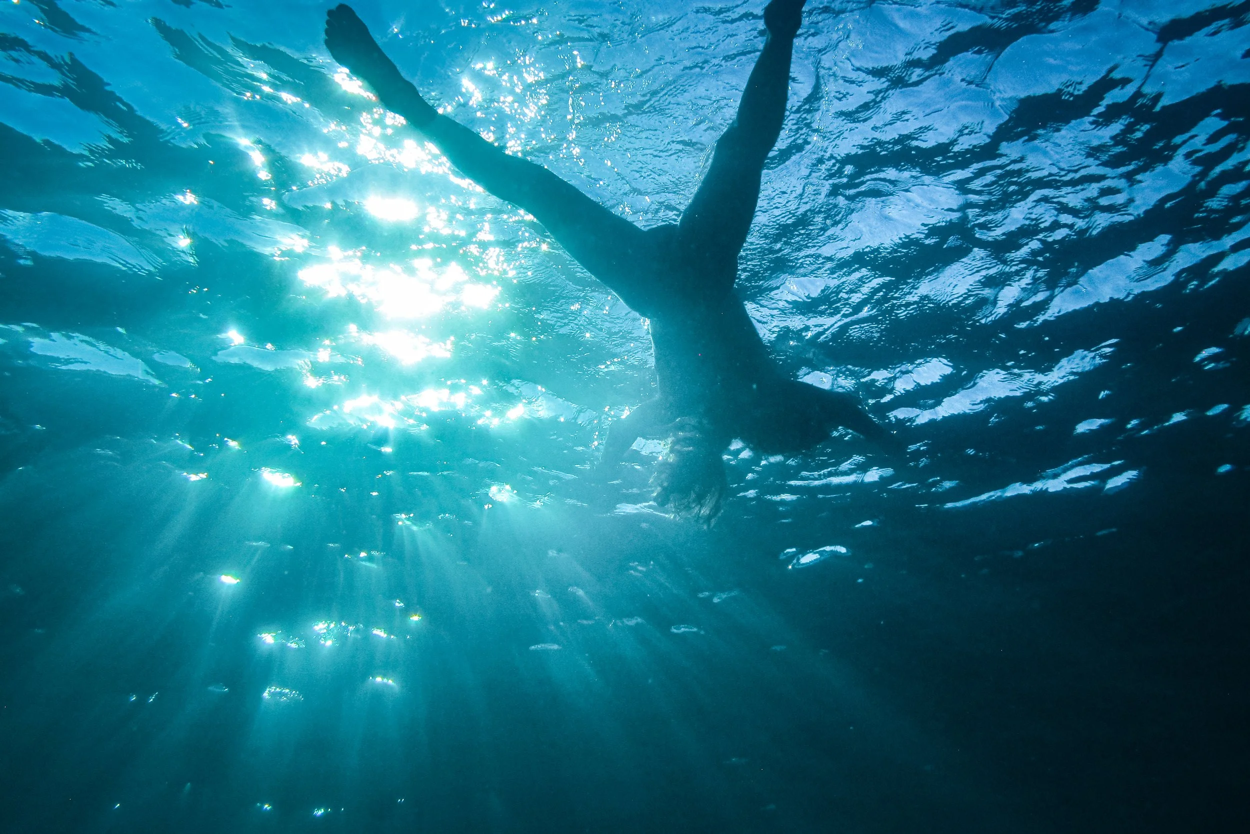 Person swimming underwater in a pool with sunlight reflecting off the water's surface.